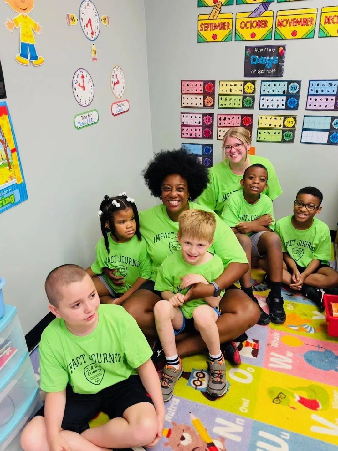 Group of children and two teachers sitting on the colorful classroom carpet, smiling for a photo. The classroom has educational posters, clocks, and calendar charts on the walls.