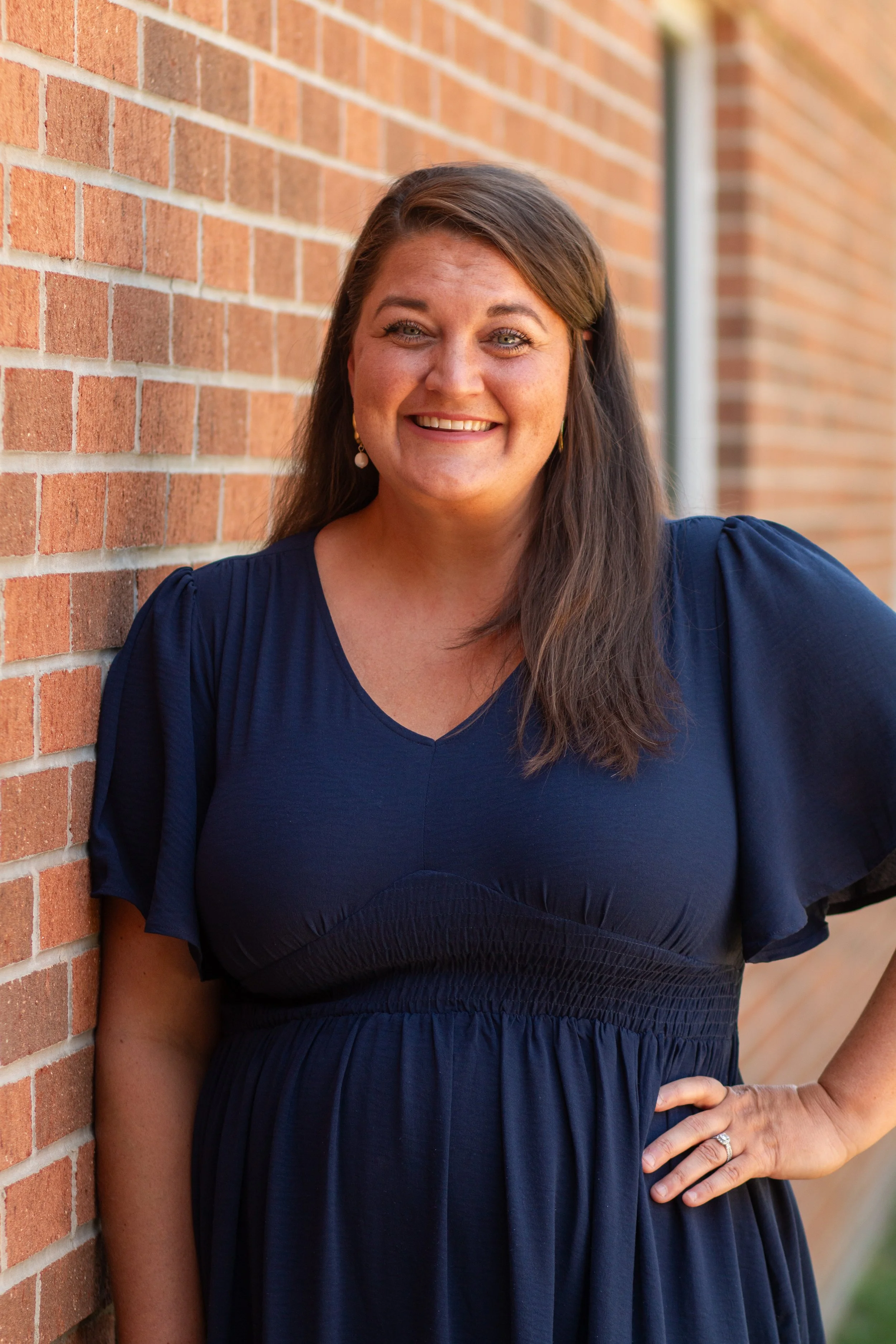 A woman with long brown hair and light skin, wearing a navy blue dress, standing outdoors next to a brick wall, smiling at the camera.