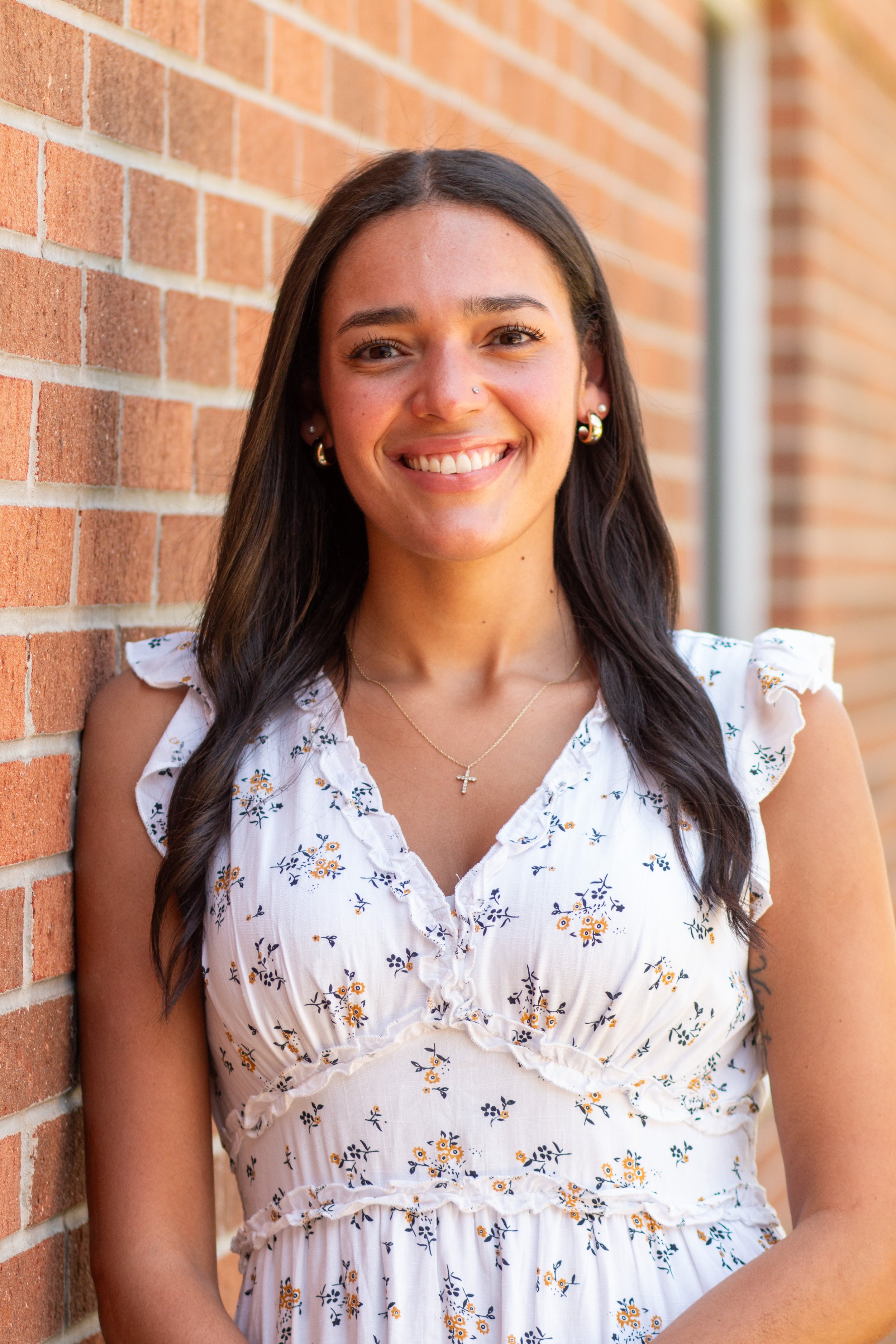 A young woman with dark brown hair, wearing a white floral dress, standing next to a brick wall with a bright smile.