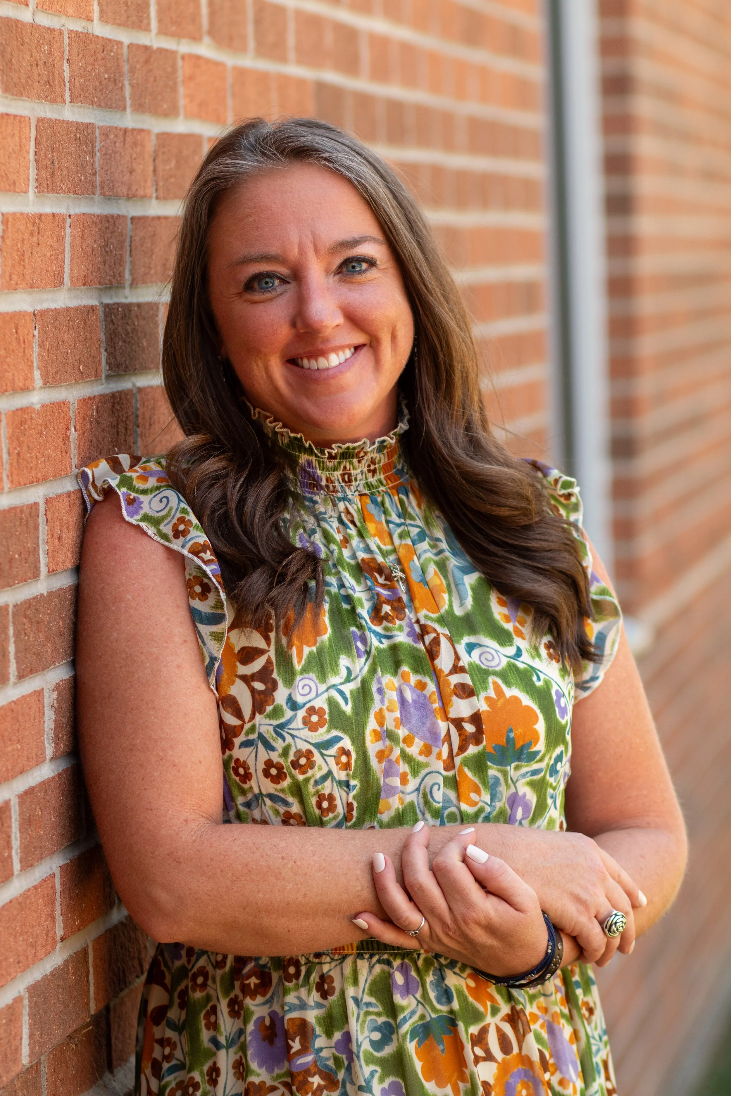 A woman with long brown hair smiling with crossed arms, standing by a brick wall during daytime.