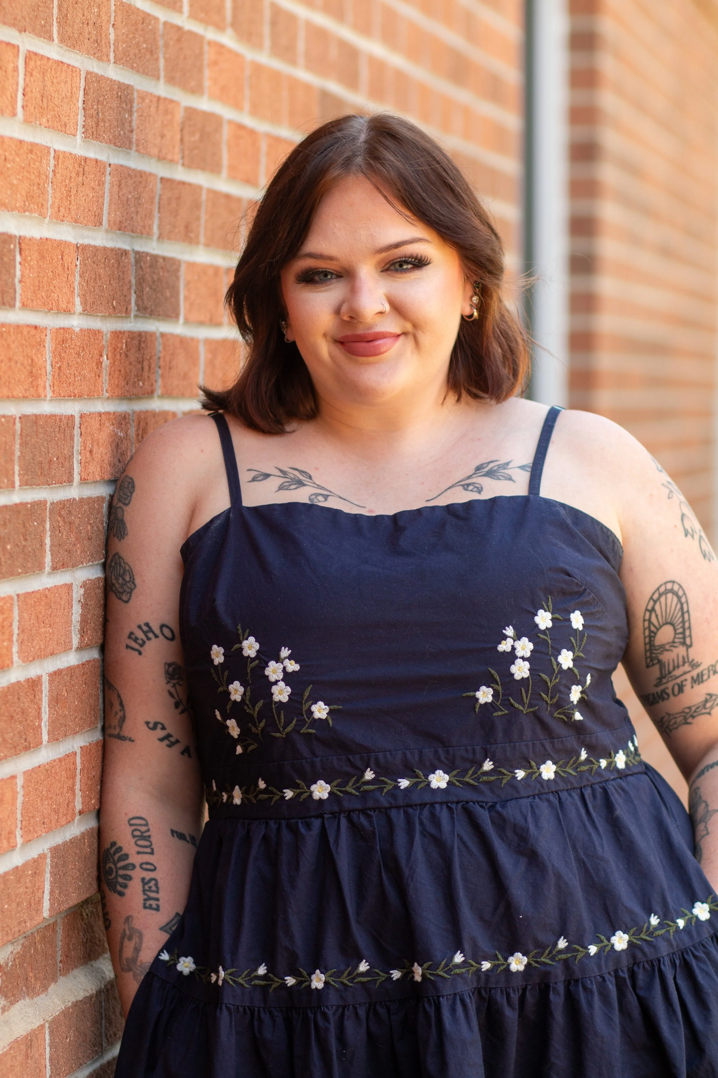 A woman with shoulder-length brown hair leaning against a brick wall, wearing a navy blue dress with floral embroidery, tattoos on her arms, earrings, and makeup.
