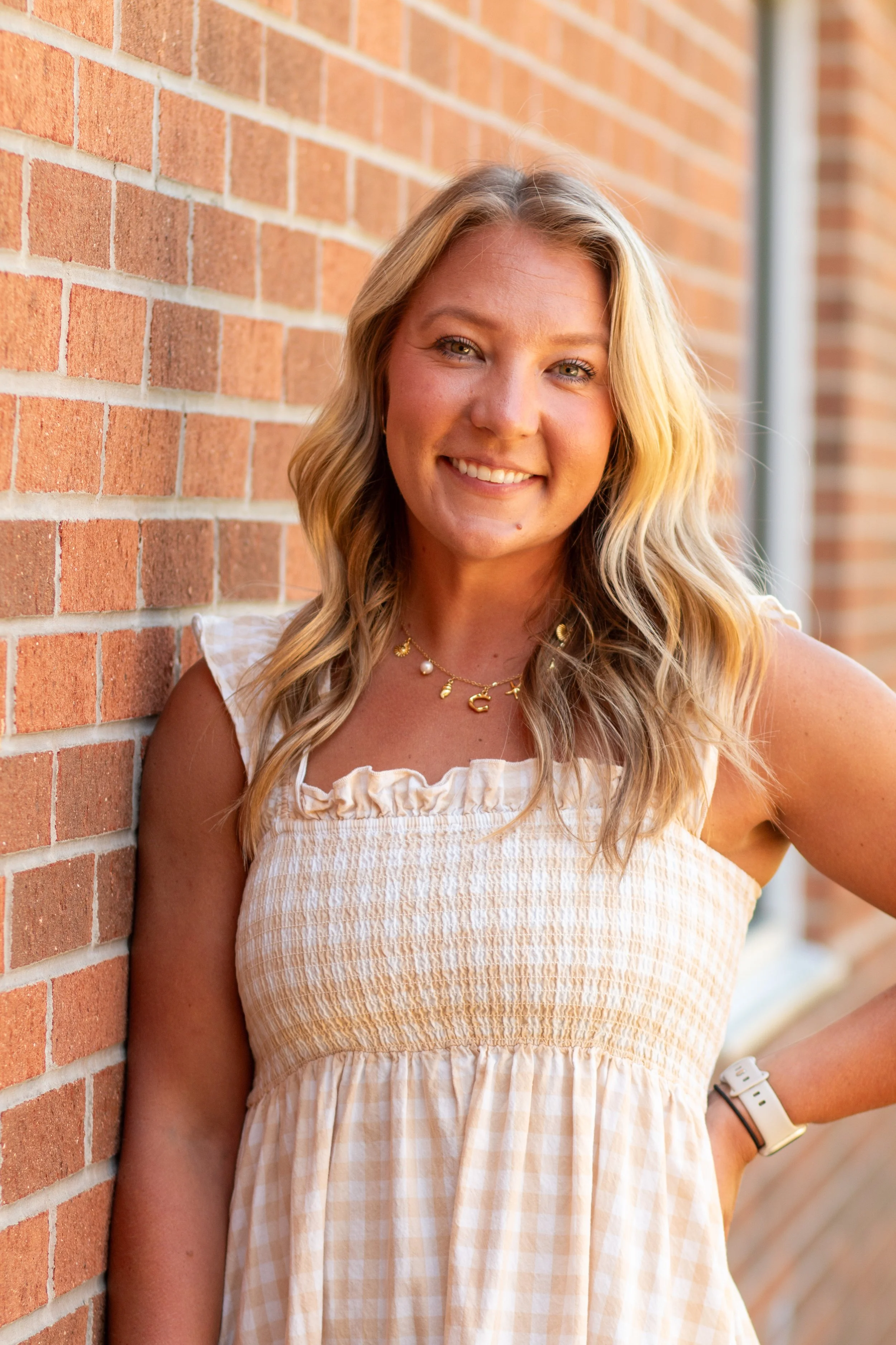 A young woman with long blonde hair standing next to a brick wall, smiling at the camera.