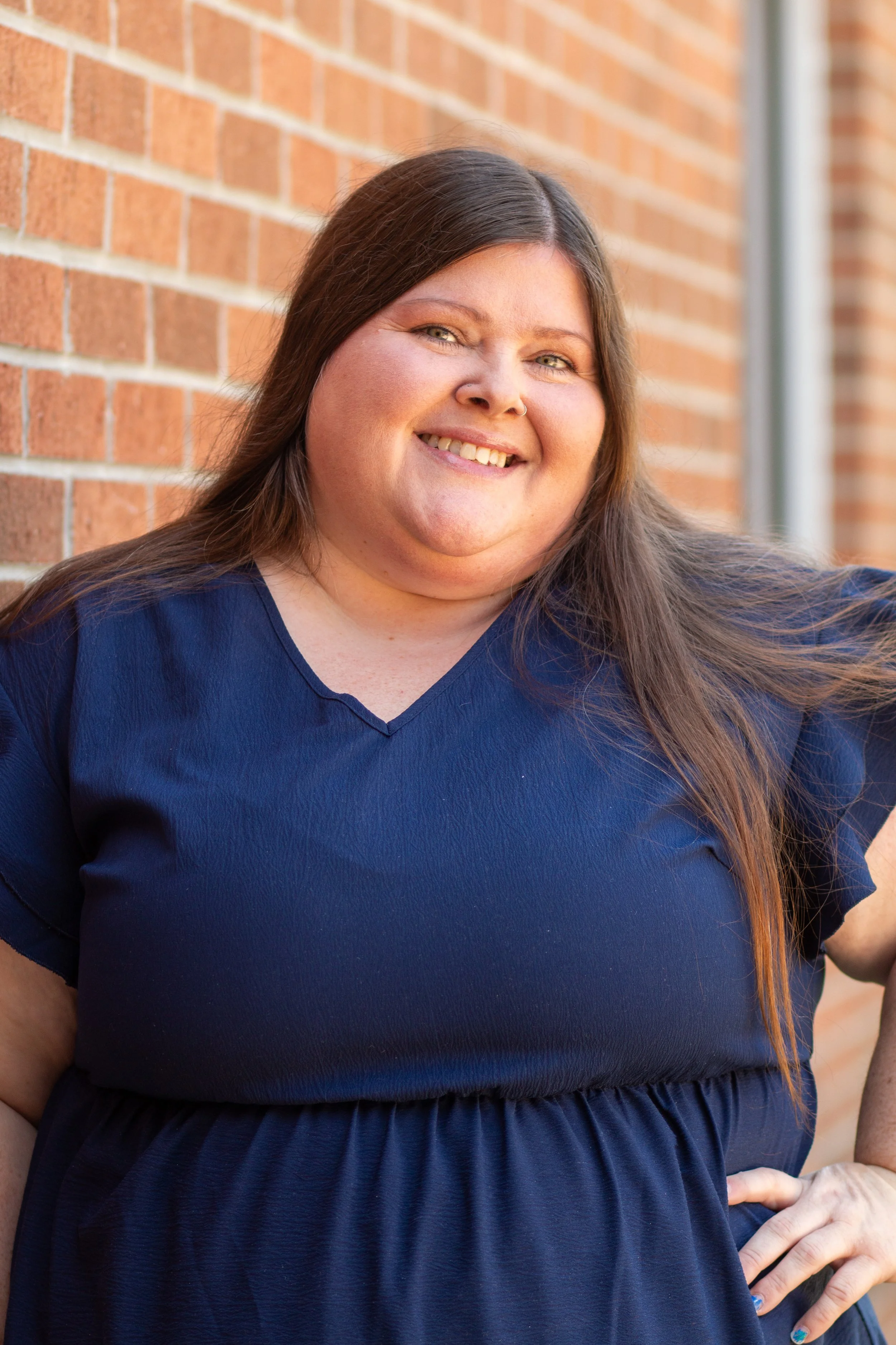 Close-up of a woman with long brown hair, wearing a navy blue dress, smiling, standing against a brick wall background.