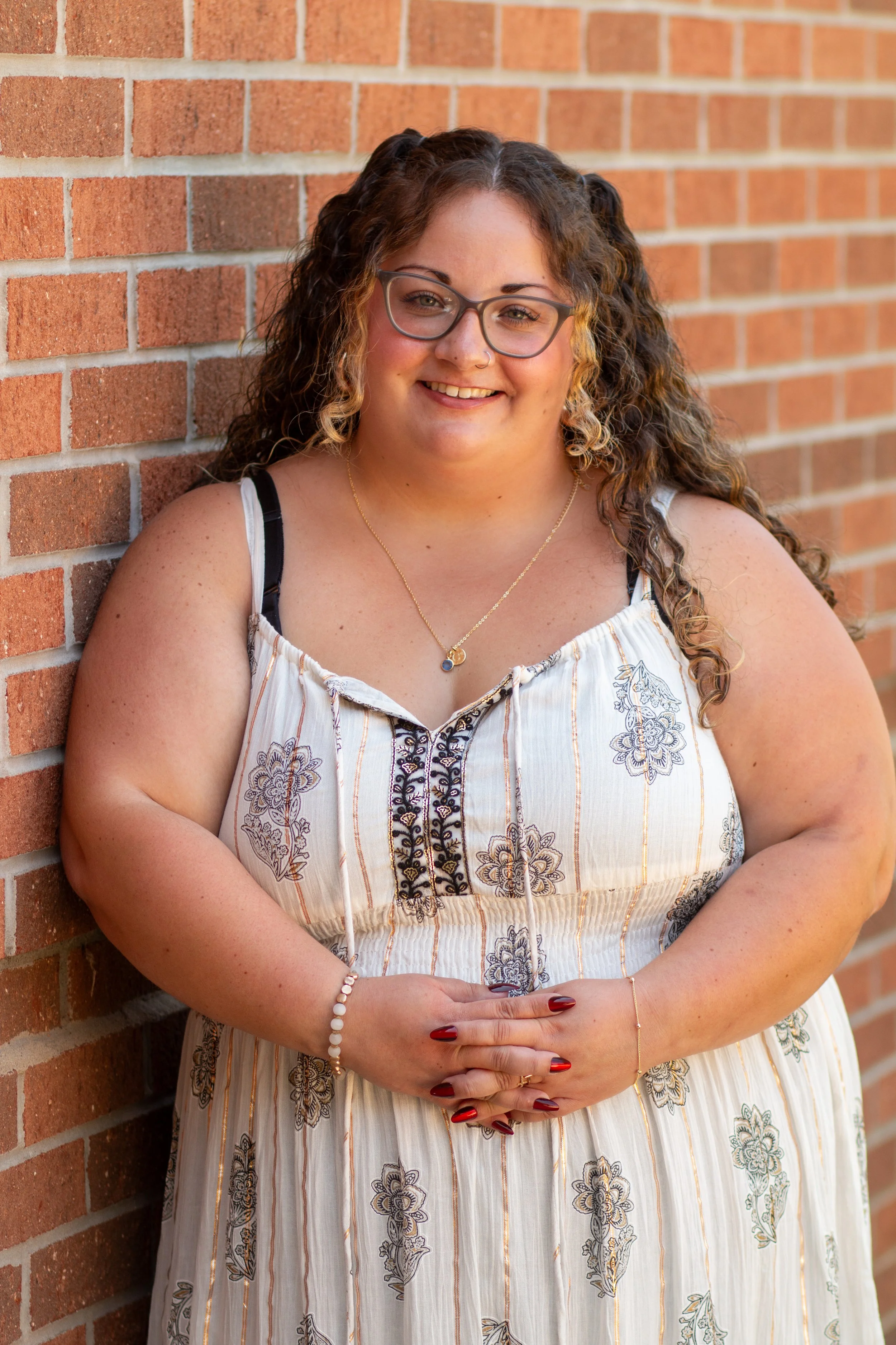 A young woman with curly brown hair and glasses, wearing a white dress with floral patterns, standing against a brick wall, smiling and clasping her hands in front of her.