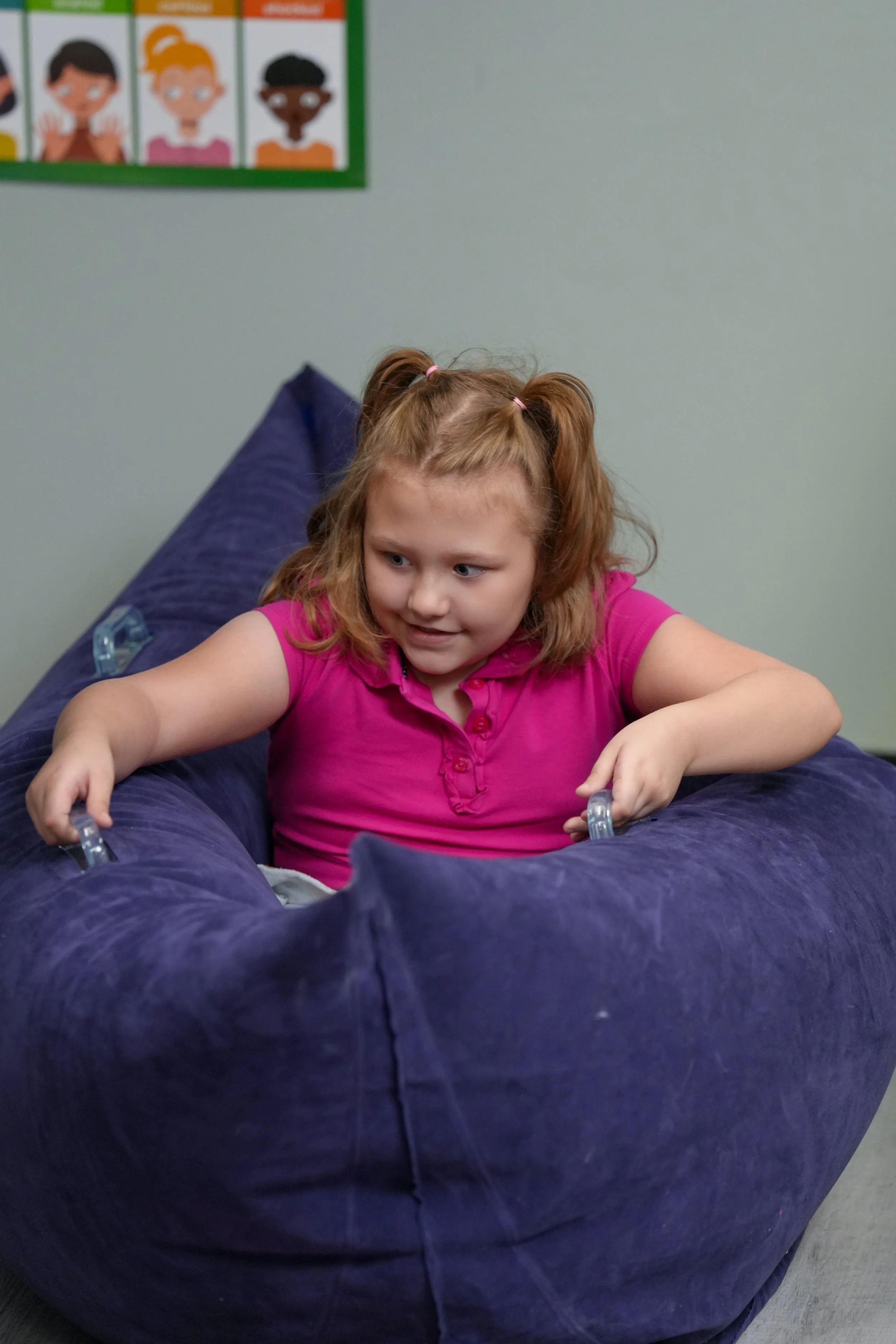 A young girl with red hair in pigtails wearing a pink shirt, smiling and playing on a large, purple, inflatable object in a classroom.