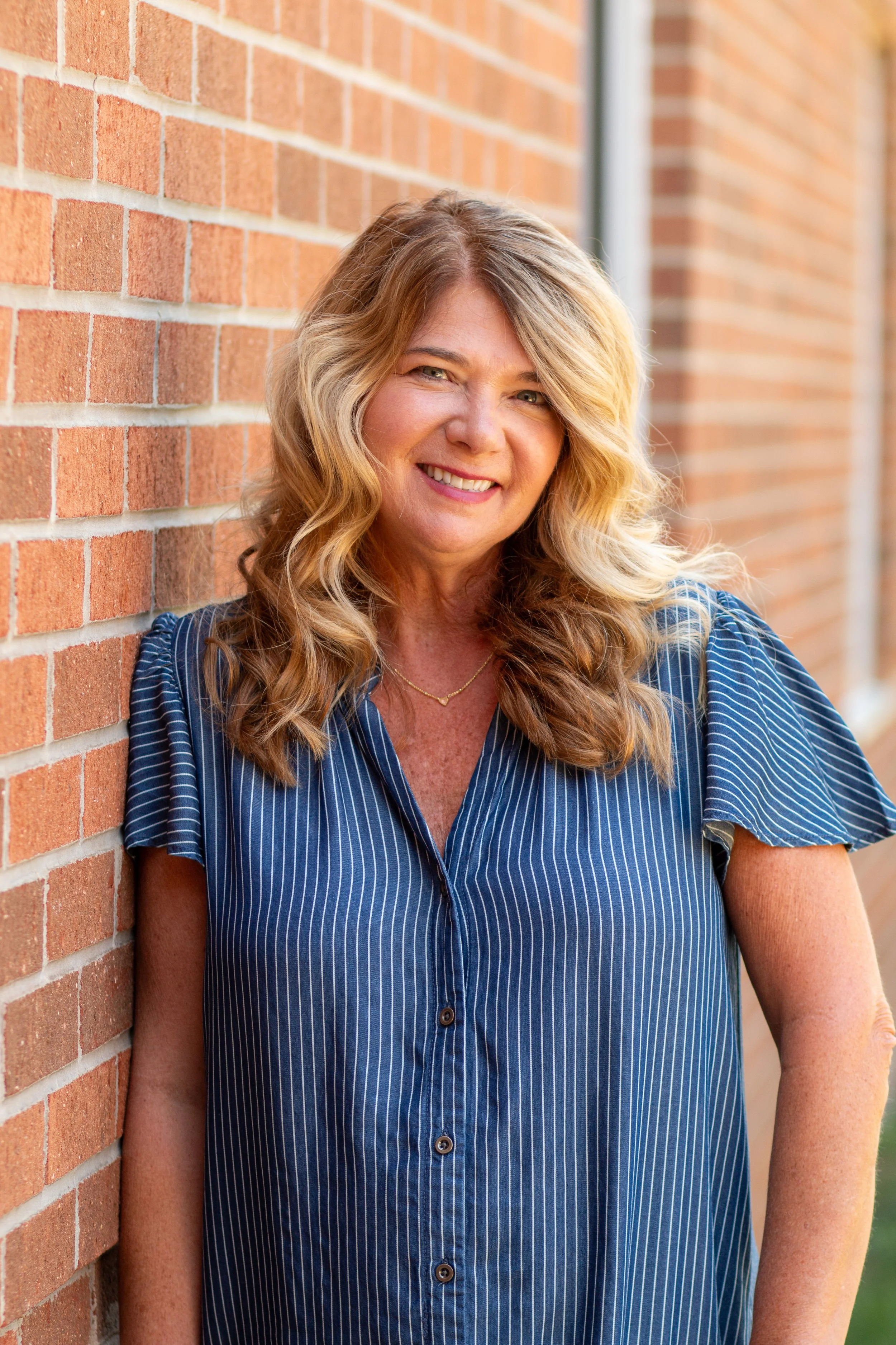 A woman with long, wavy blond hair smiling while leaning against a brick wall, wearing a blue pinstripe shirt.