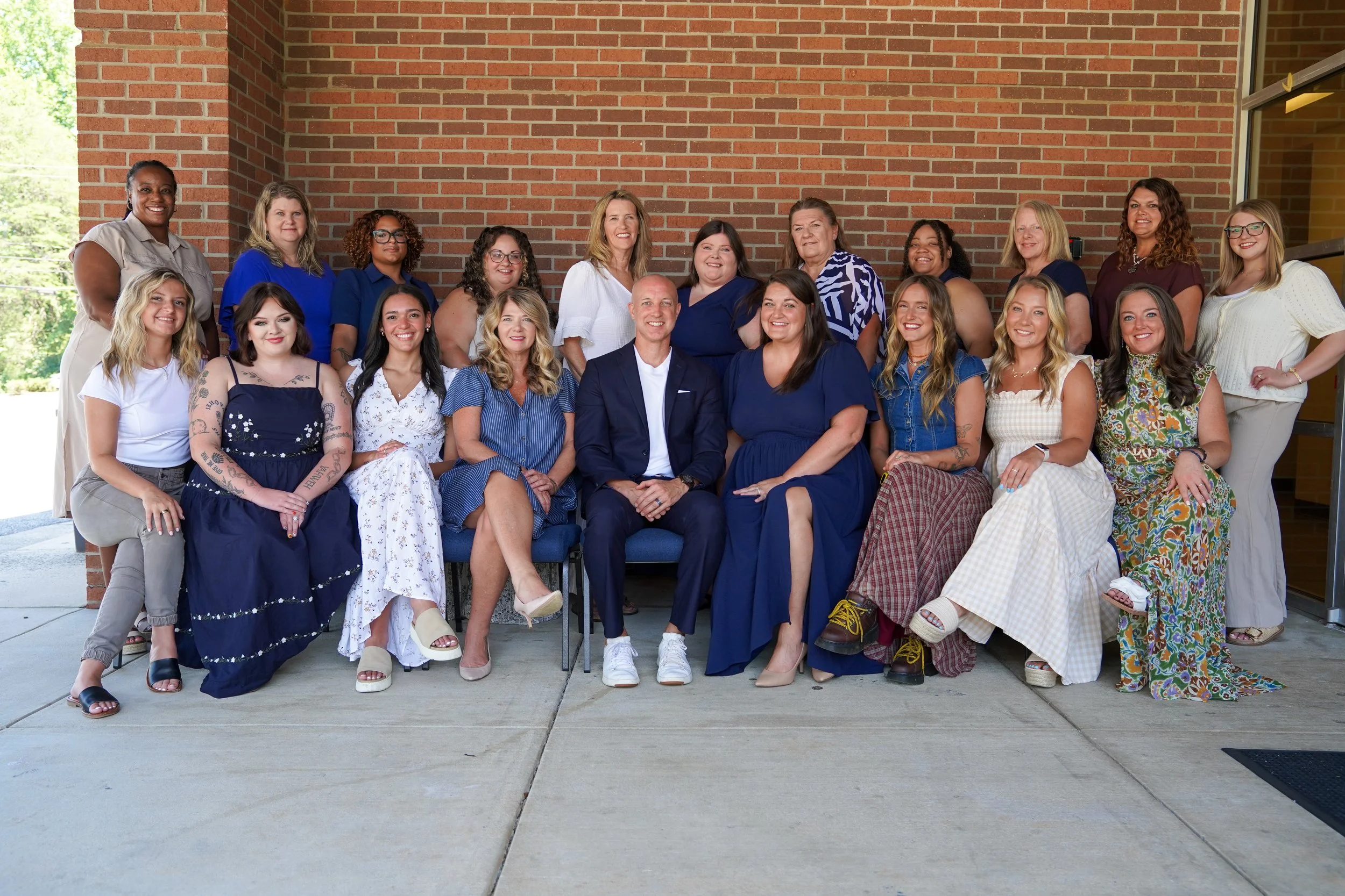 Group of diverse women and one man posing for a photo outside a building with a brick wall in the background. They are smiling and sitting and standing in a semi-organized manner.