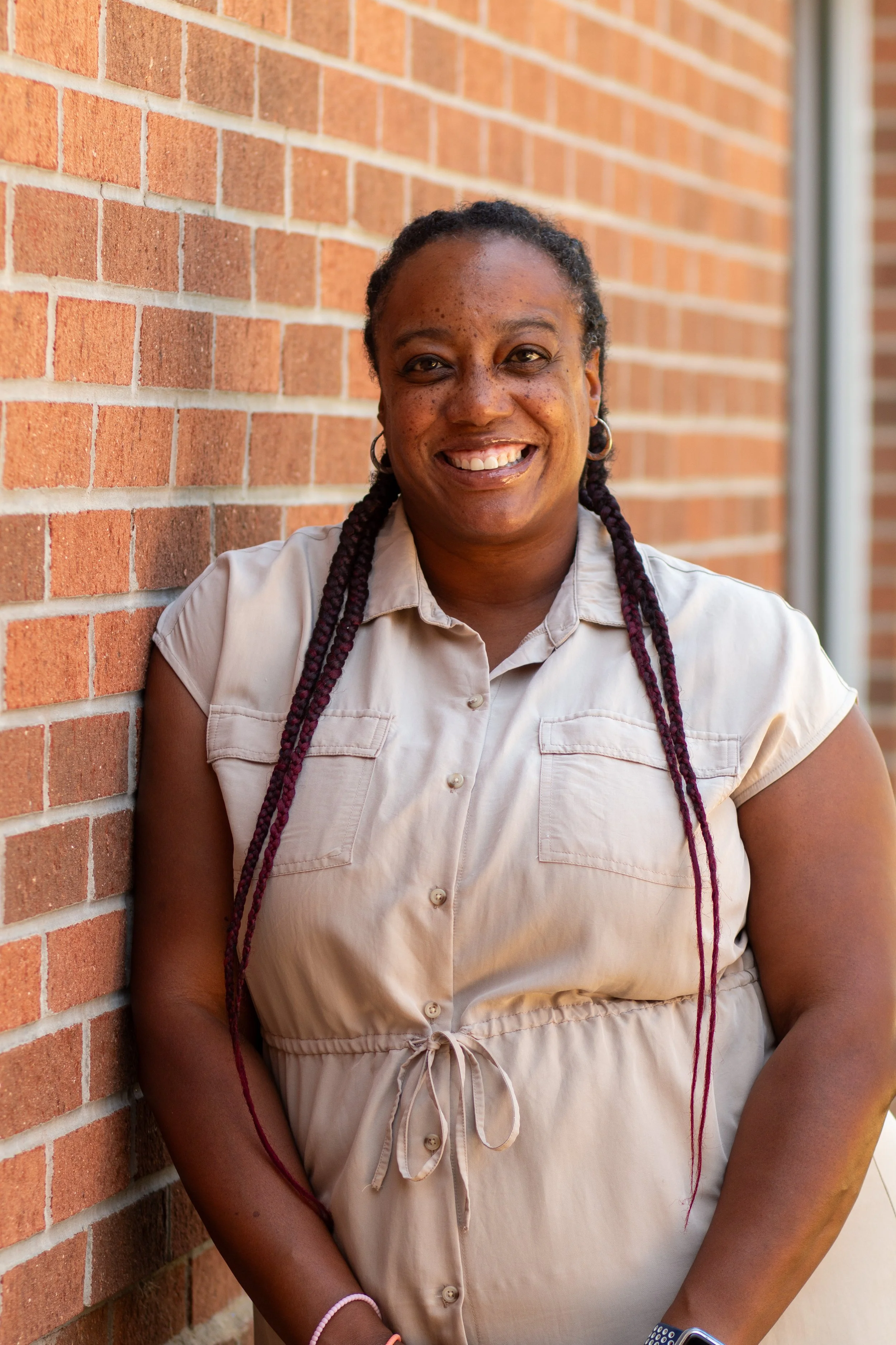 A woman with braided hair and earrings smiling while leaning against a brick wall.
