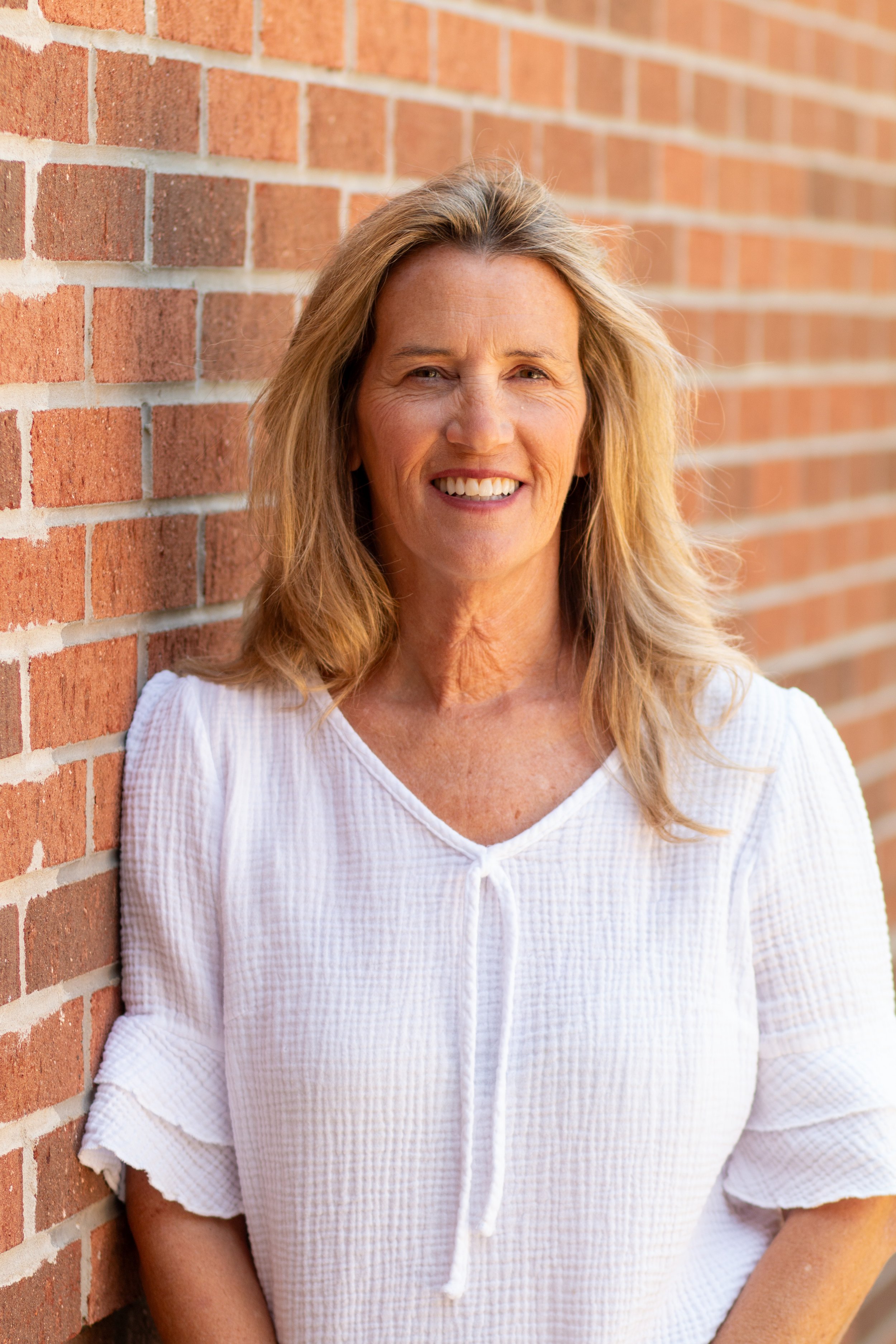A middle-aged woman with blonde hair, smiling, standing against a red brick wall.