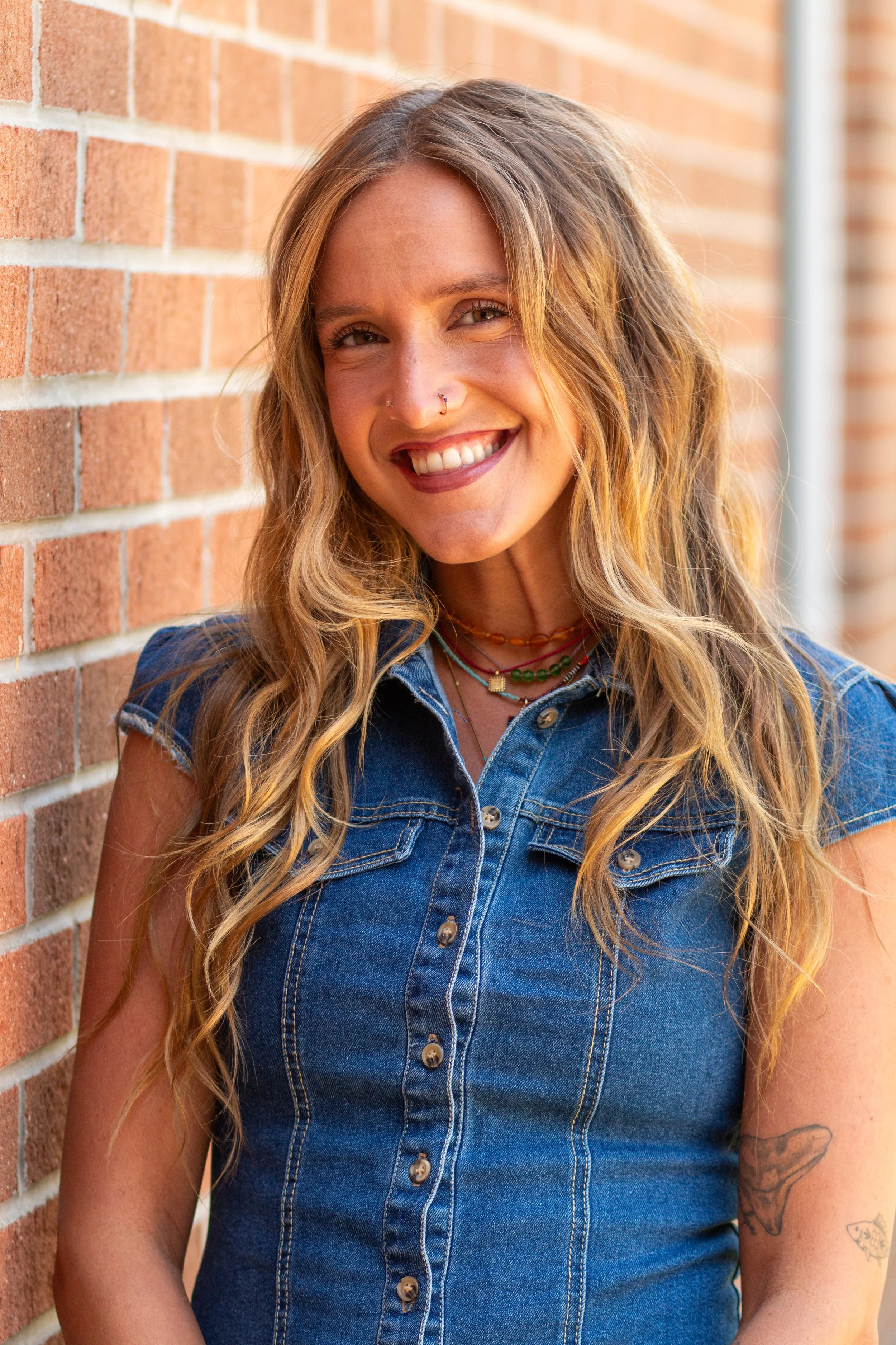 A smiling woman with long wavy hair, wearing a sleeveless denim shirt and multiple necklaces, standing outdoors against a brick wall.