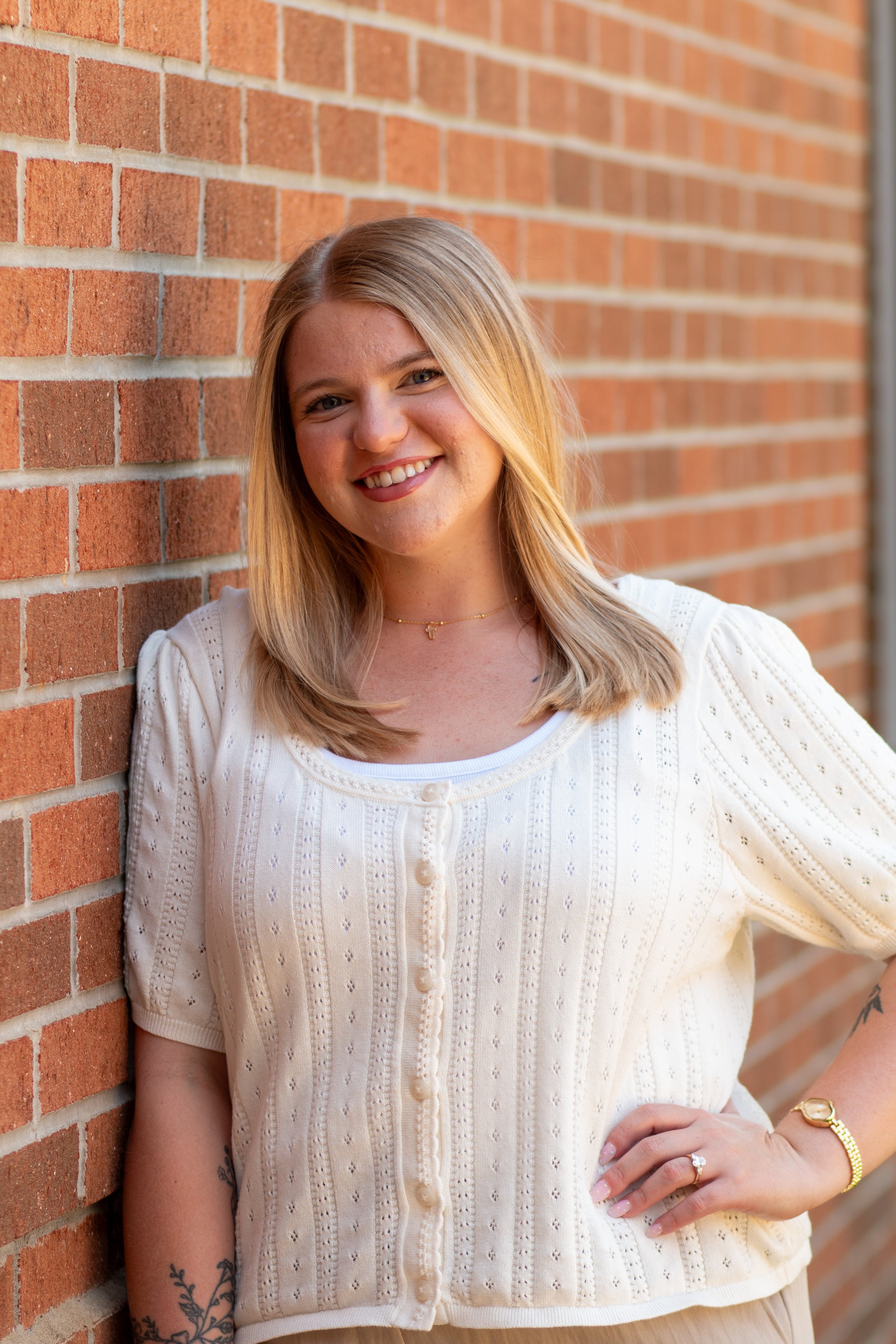 A woman with blonde hair smiling, standing against a brick wall, wearing a cream-colored knitted cardigan and jewelry.