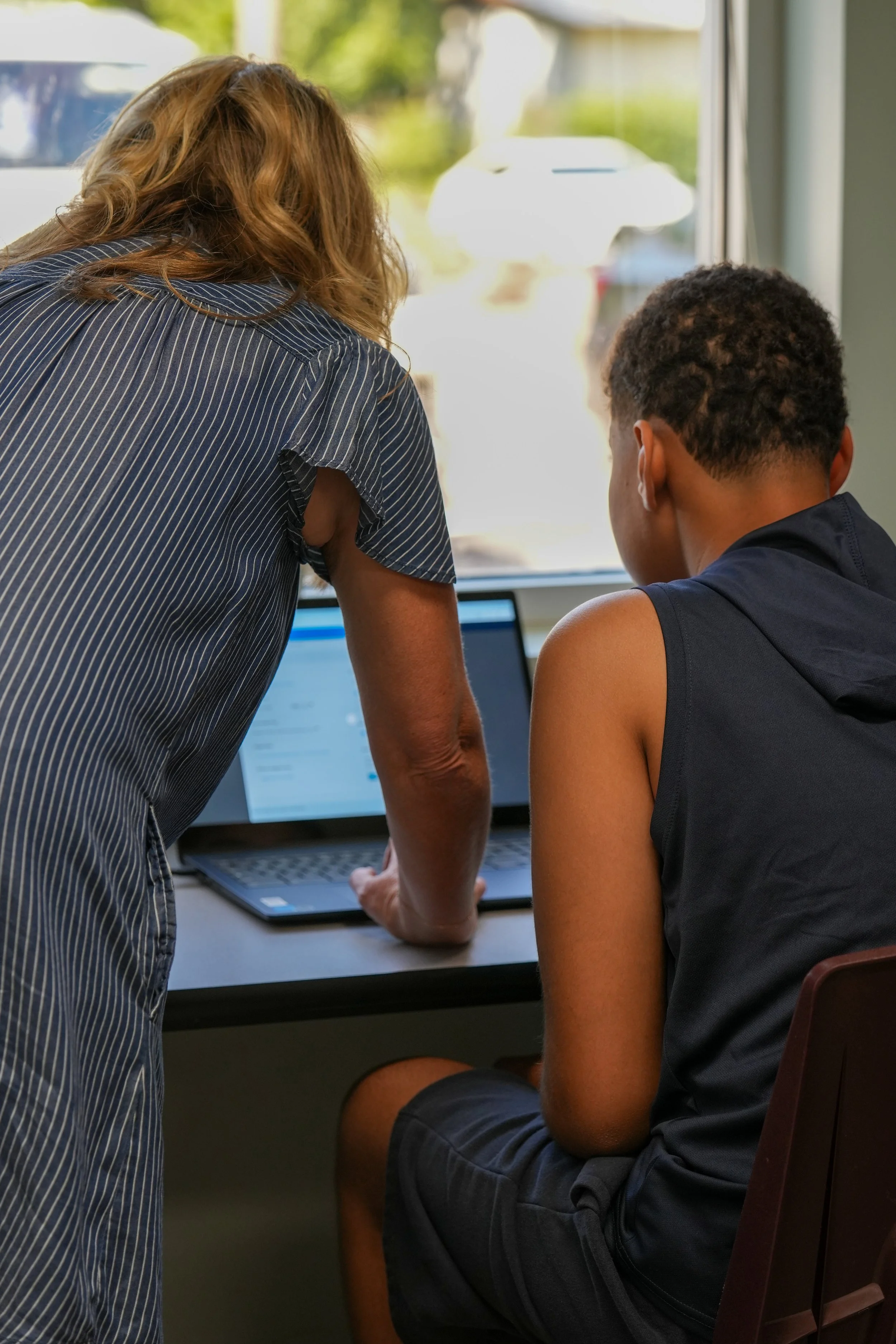 A teacher or adult woman and a male student working together on a laptop computer in a classroom or library. They are both focused on the screen, with the teacher leaning over to assist the student.