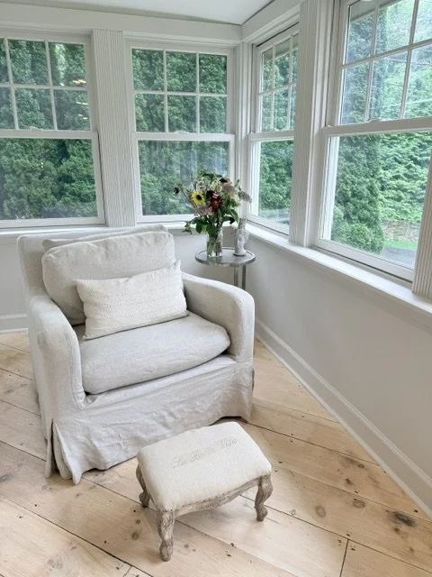 Sunroom with a beige armchair, matching pillow, white wooden flooring, and large windows showing green trees outside, with a small round side table holding a vase of colorful flowers.