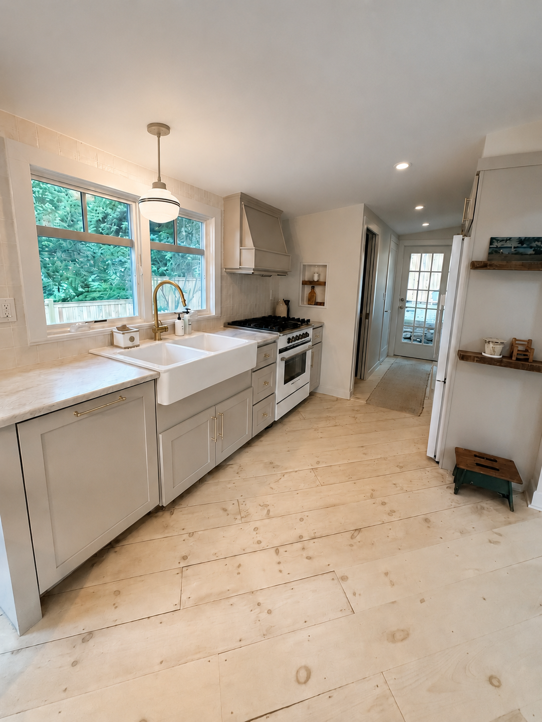 Bright kitchen with beige cabinets, white farmhouse sink, gold faucet, gas stove, light wood flooring, two large windows showing greenery, and a glass door leading outside.