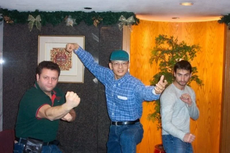 Three men striking martial arts poses indoors. The man on the left is wearing a green shirt with red accents, the man in the middle is wearing glasses and a blue plaid shirt with a blue cap, and the man on the right is wearing a light gray sweater. T