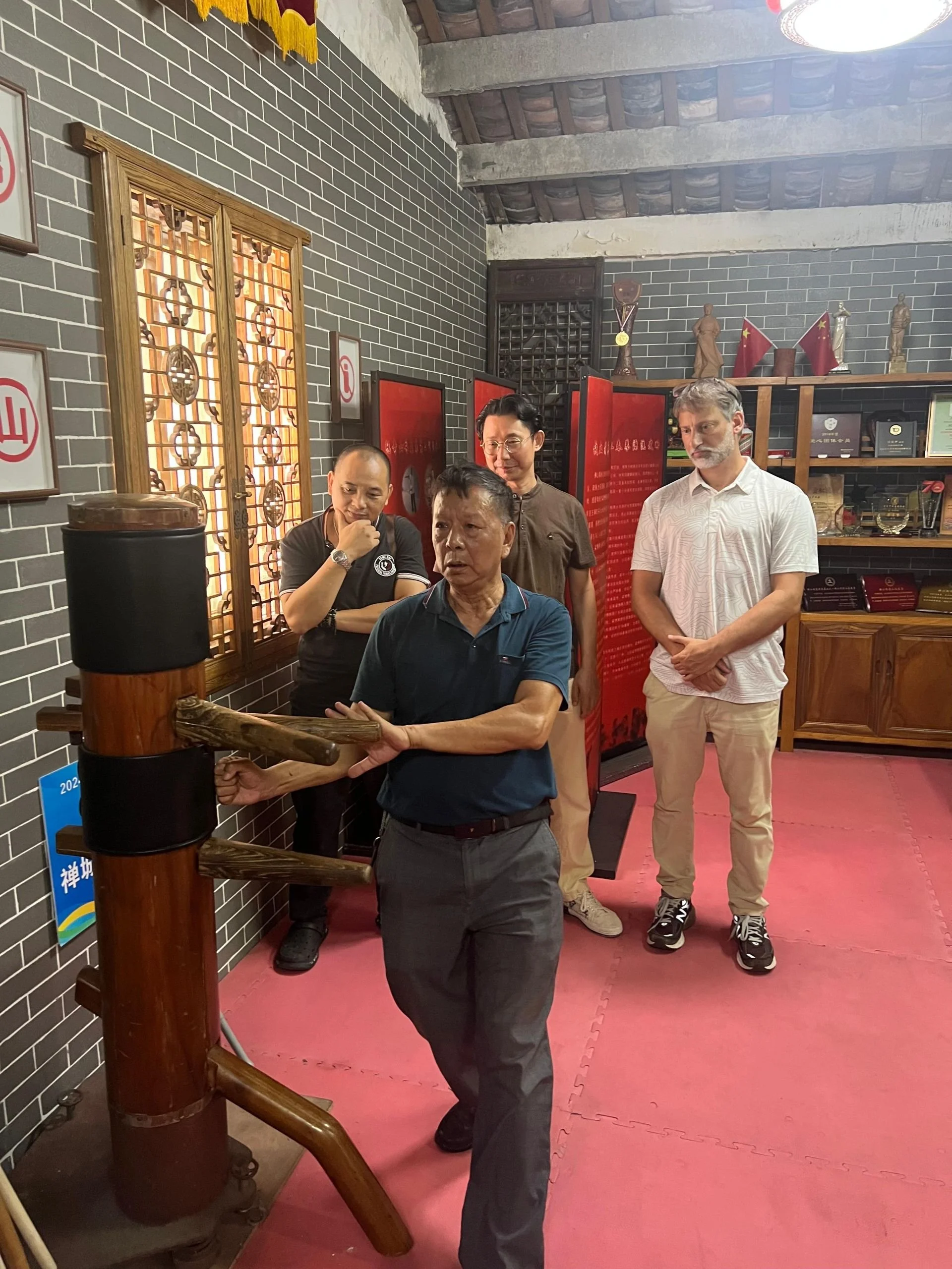 Five people stand around a punching bag inside a room with black brick walls and wooden ceiling beams. The person in the foreground is in action, punching the bag. The others observe, with two men and two women among them. Shelves with awards and fla