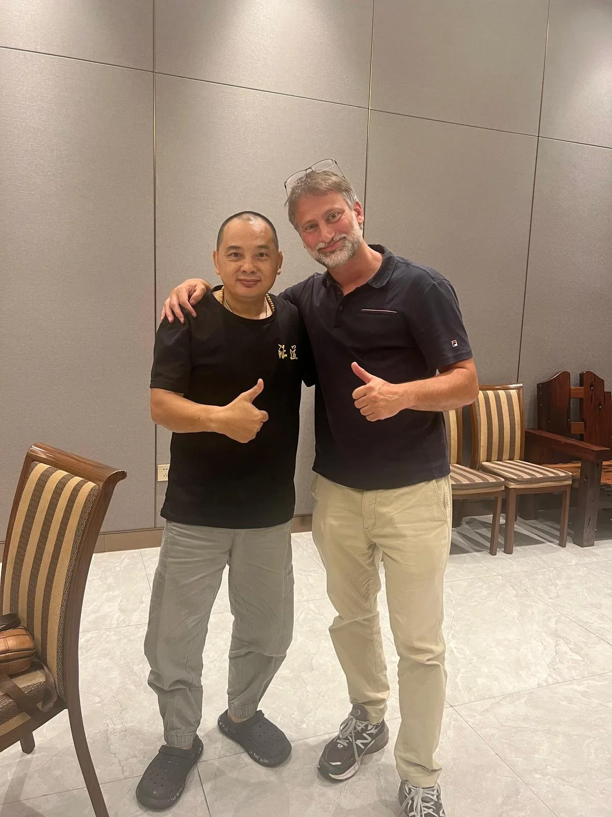 Two men standing together indoors, giving a thumbs-up. The man on the left is wearing a black t-shirt and light gray pants, and the man on the right is wearing a black polo shirt and beige pants. They are smiling and have their arms around each other