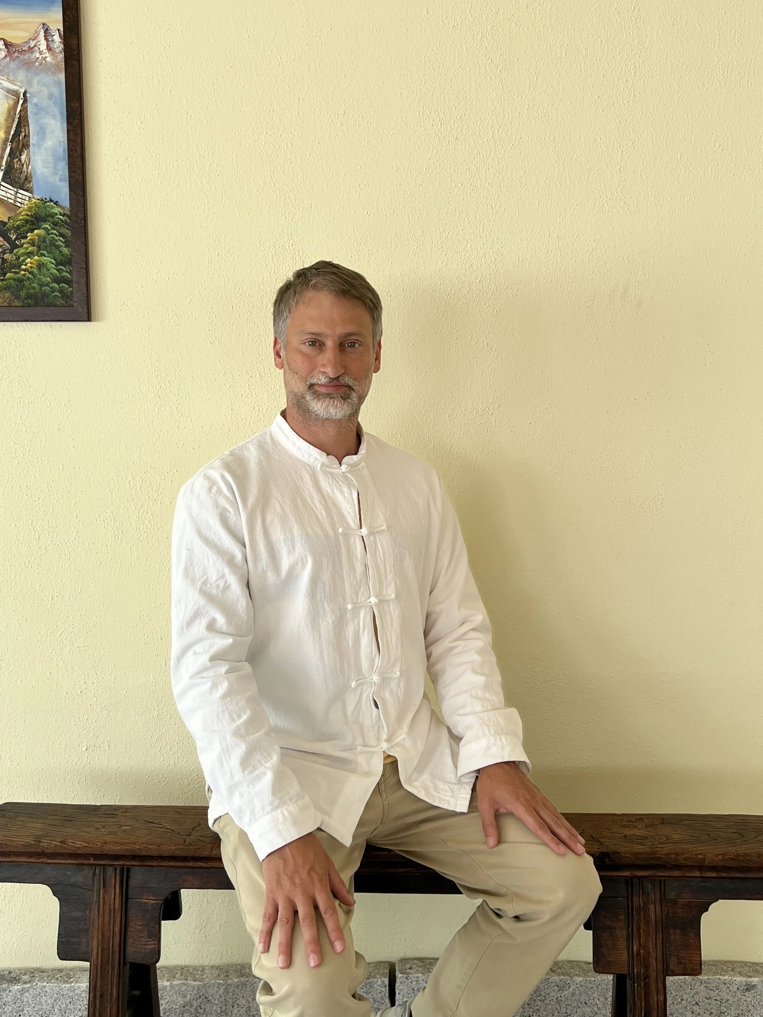 A man with gray hair and beard wearing a white traditional Chinese shirt and beige pants, sitting on a wooden bench against a yellow wall, with a framed painting partially visible on the wall.