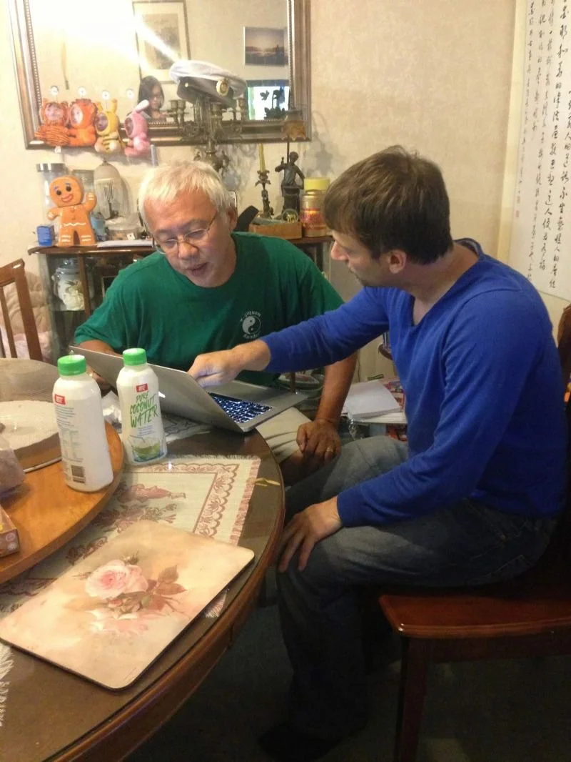 Two men are sitting at a dining table looking at a tablet together in a warmly decorated room with various toys and decorative items on a sideboard in the background.