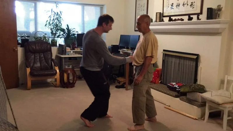 Two men practicing martial arts or self-defense moves in a living room, facing each other barefoot.
