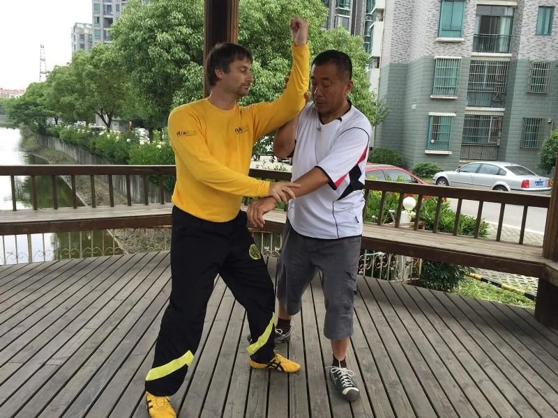 Two men practicing martial arts on a wooden deck, with city buildings and parking lot in the background.