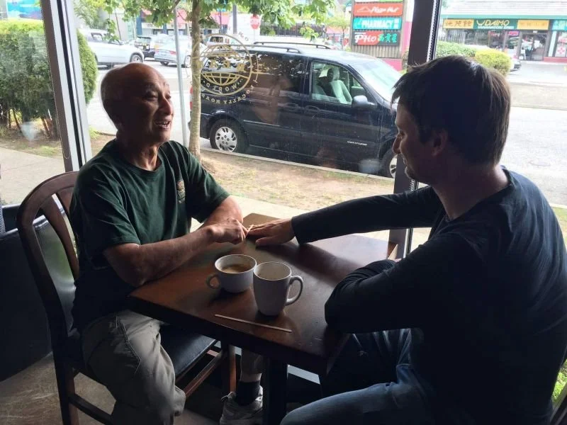 An elderly man and a young man are sitting and holding hands at a table inside a coffee shop, with cups of coffee on the table. They are near a large window with a view of parked cars and buildings outside.