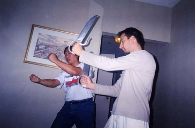 Two men holding swords in a pretend sword fight inside a room with a framed picture on the wall.