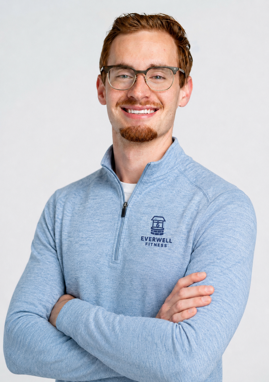 A smiling man with red hair, glasses, and a goatee, wearing a light blue zip-up sports jacket with the Everwell Fitness logo on it, standing against a plain light background.