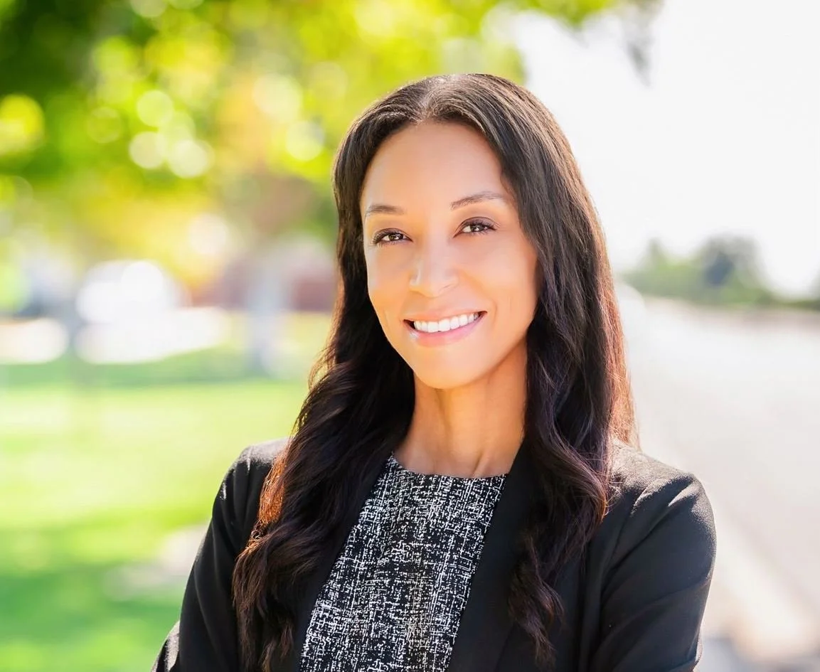 A smiling woman with long dark hair, wearing a black blazer and patterned blouse, standing outdoors on a sunny day with blurred greenery in the background.