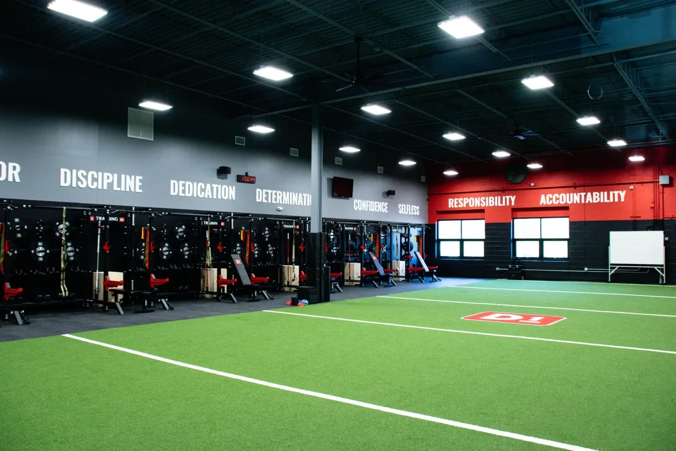 Indoor gym with black workout equipment and sports training stations. The wall displays motivational words such as 'Discipline,' 'Dedication,' 'Confidence,' 'Responsibility,' and 'Accountability.' The floor has artificial green turf with white lines and a red logo. There are two windows on the back wall.