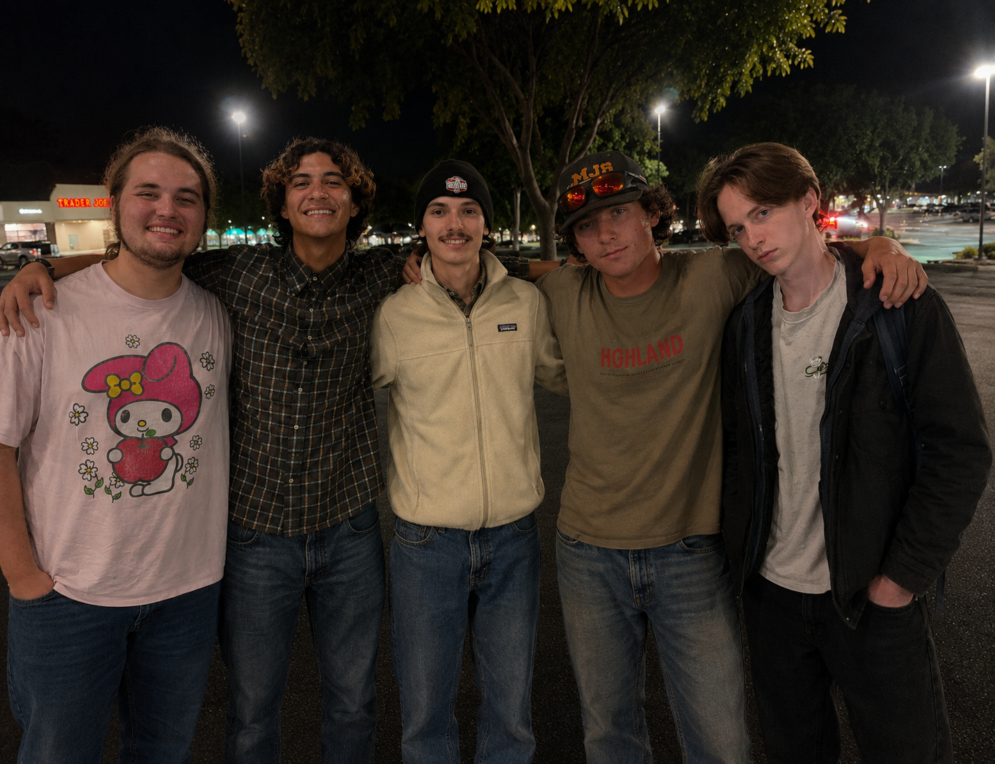 Five young men standing side by side with arms around each other's shoulders in a parking lot at night, smiling at the camera, illuminated by streetlights and store signs in the background.