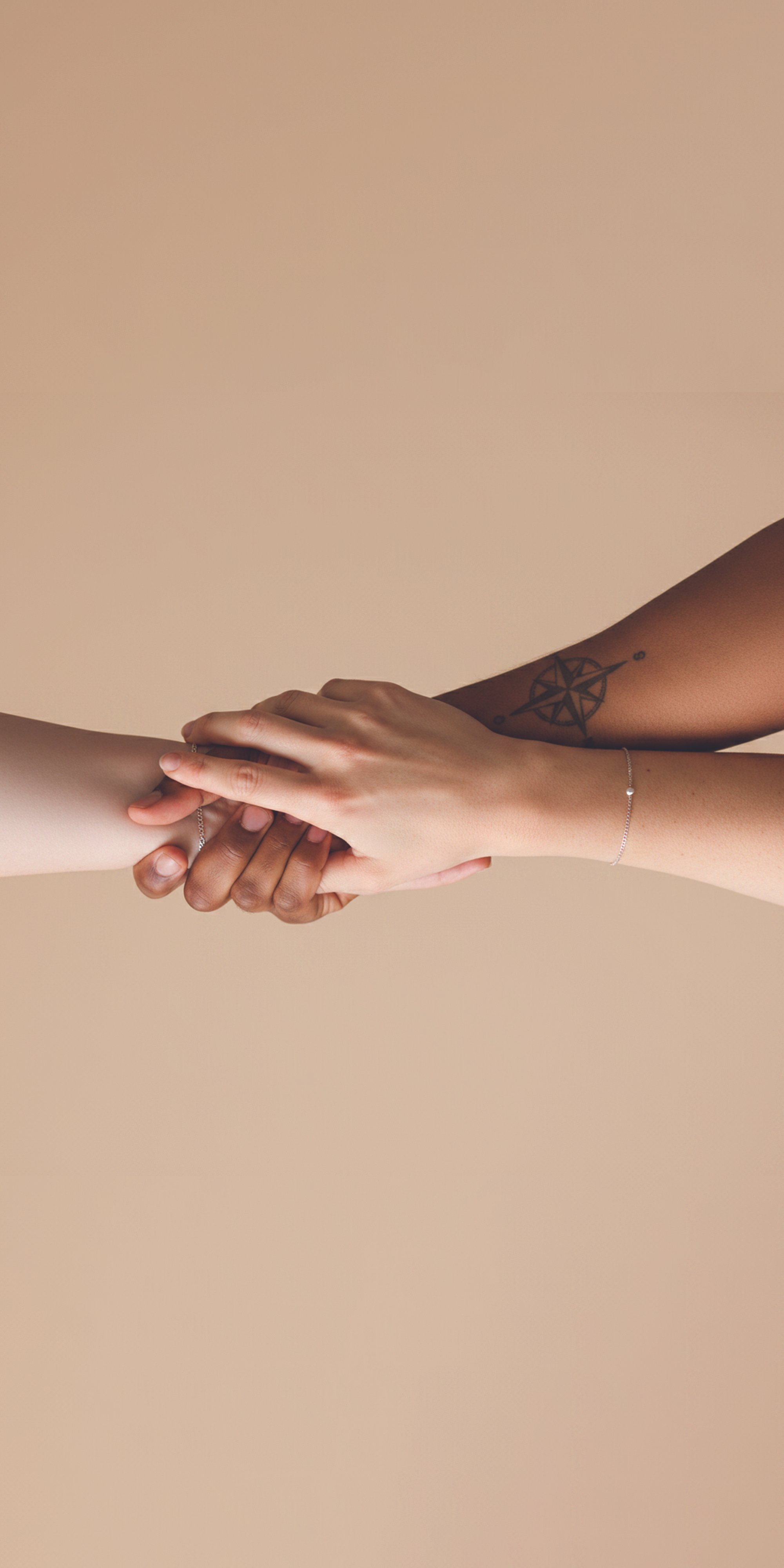 Two people holding hands against a beige background, one with dark skin and the other with light skin, wearing delicate jewelry.