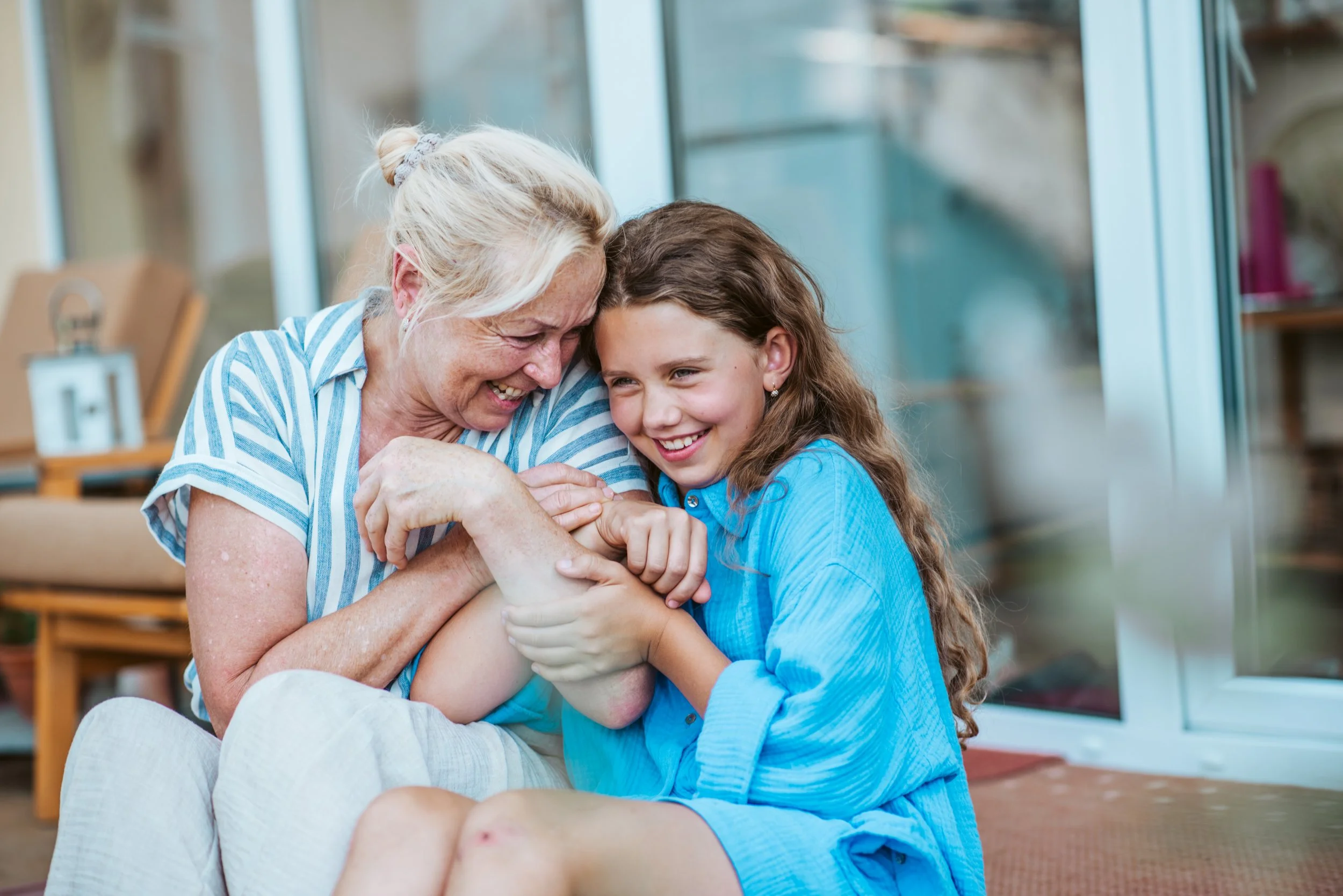 A grandmother and her granddaughter smiling and embracing each other indoors, with the grandmother wearing a blue and white striped shirt and the granddaughter in a bright blue shirt.