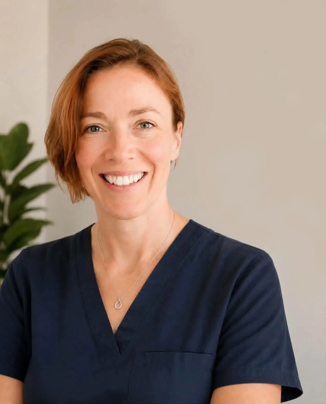 Portrait of a woman with short red hair smiling, wearing a navy blue top and a silver necklace, standing in front of a light-colored wall with a green plant in the background.