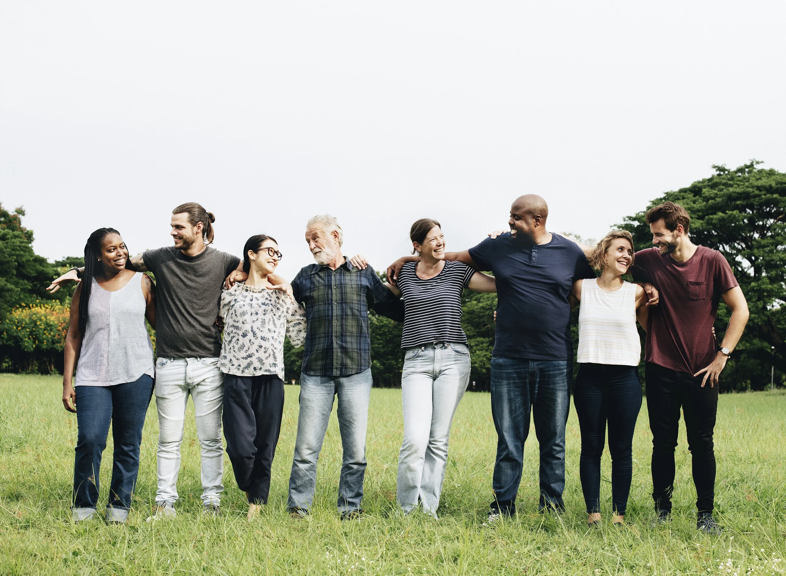 Group of eight diverse friends standing in a park with their arms around each other, smiling and laughing on a cloudy day.