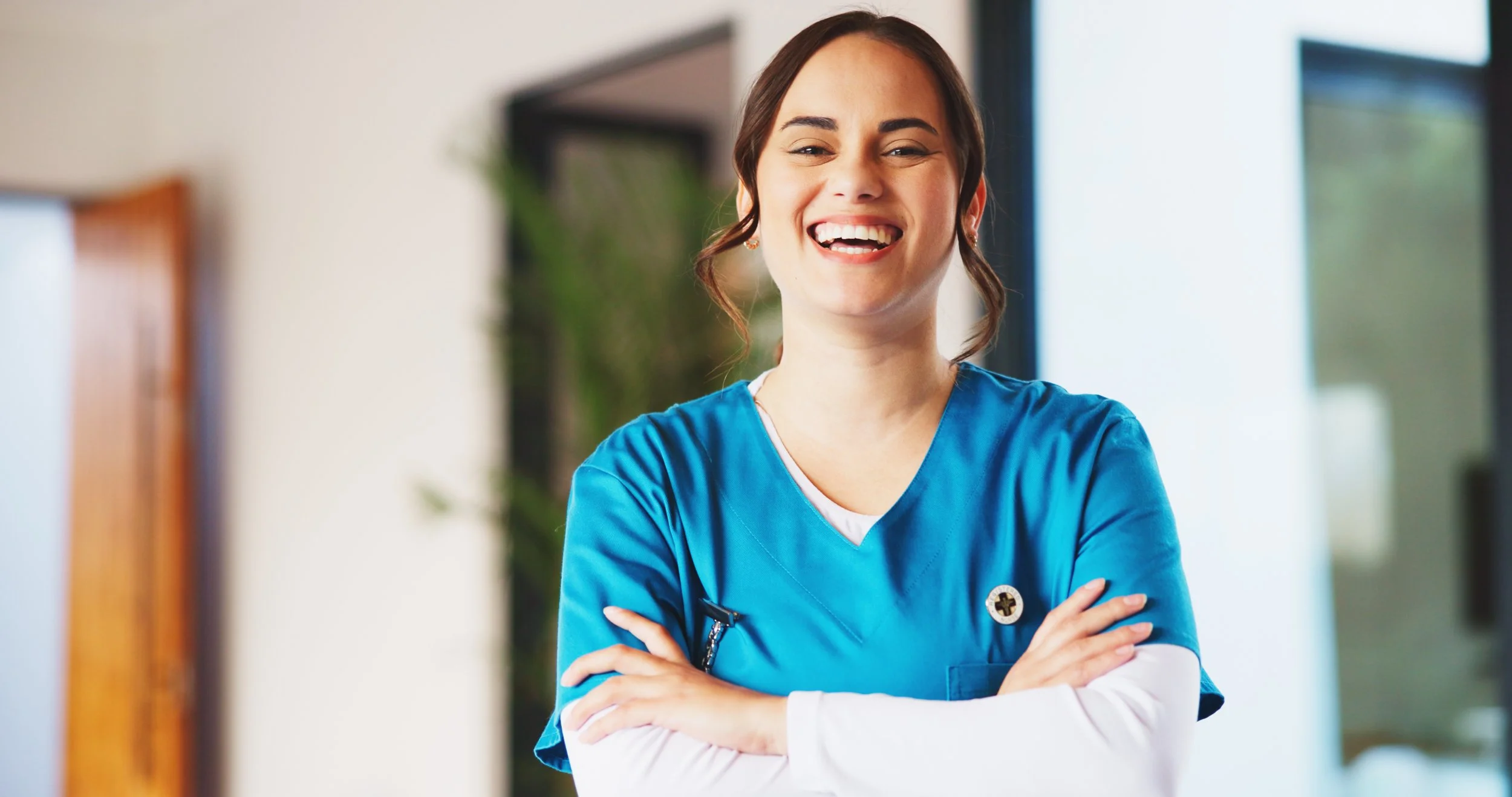 A smiling female nurse in blue scrubs with arms crossed, standing in a well-lit modern healthcare setting.