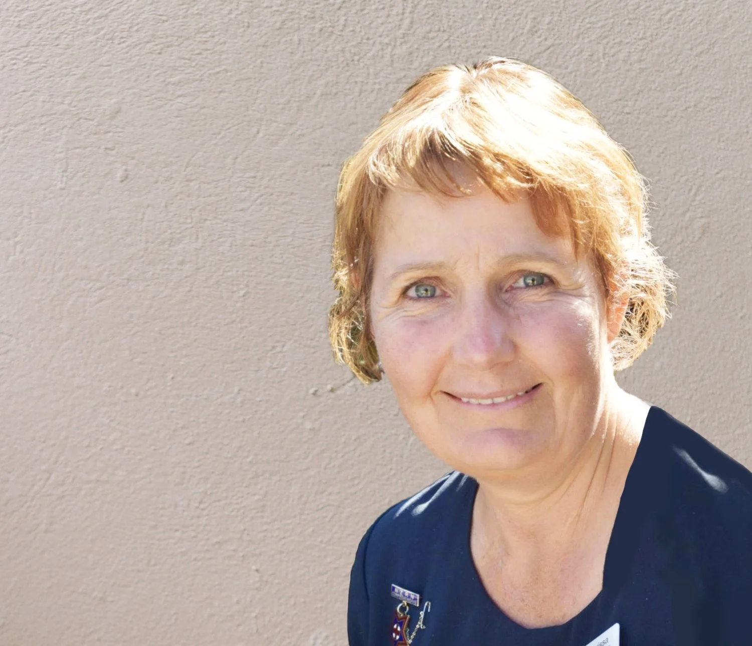 A woman with short, reddish-blonde hair and light skin, smiling and wearing a dark shirt, standing in front of a beige textured wall.