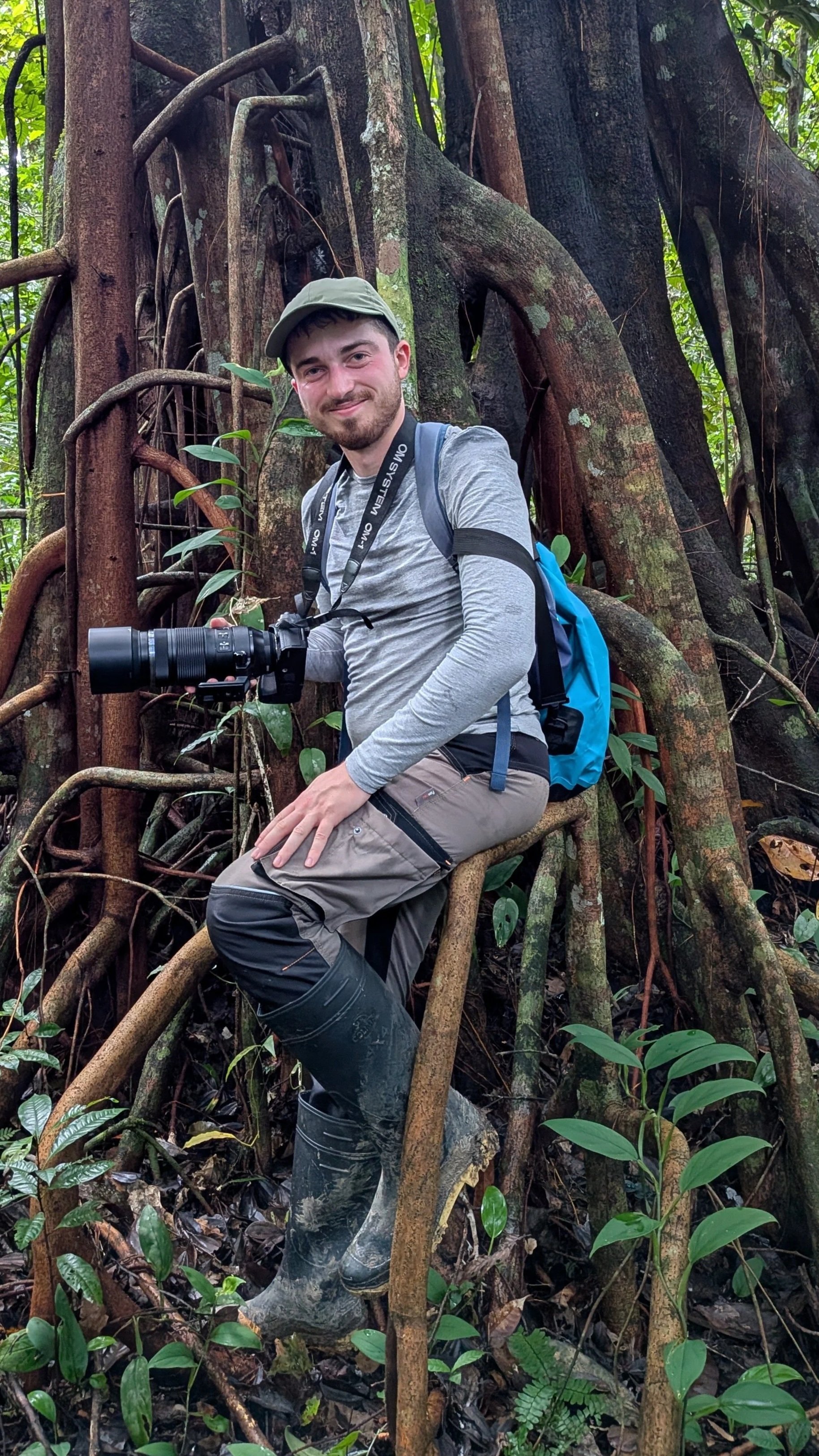 A man with a beard, wearing a grey long-sleeve shirt, grey cap, and muddy boots, sitting on a large root in a lush jungle, holding a DSLR camera with a telephoto lens, and smiling at the camera.