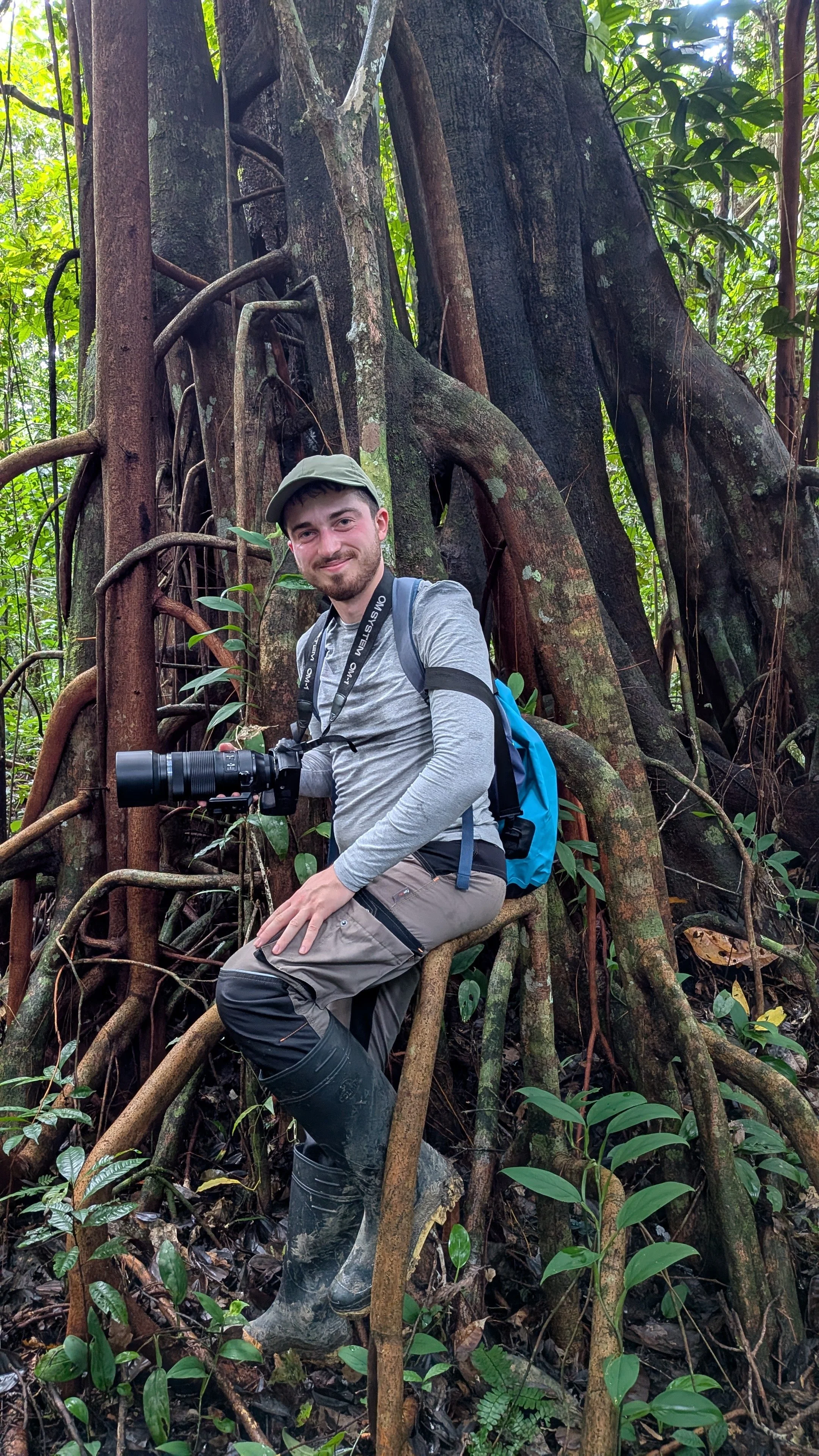 A man in outdoor gear, including a gray long-sleeve shirt, gray pants, and rubber boots, is sitting on a root in a dense jungle, holding a camera with a large telephoto lens around his neck. He is smiling and wearing a light gray cap, with a blue backpack on his back. The surrounding environment is lush with green leaves and large tree roots, with the man positioned in front of a massive tree with thick roots and branches.
