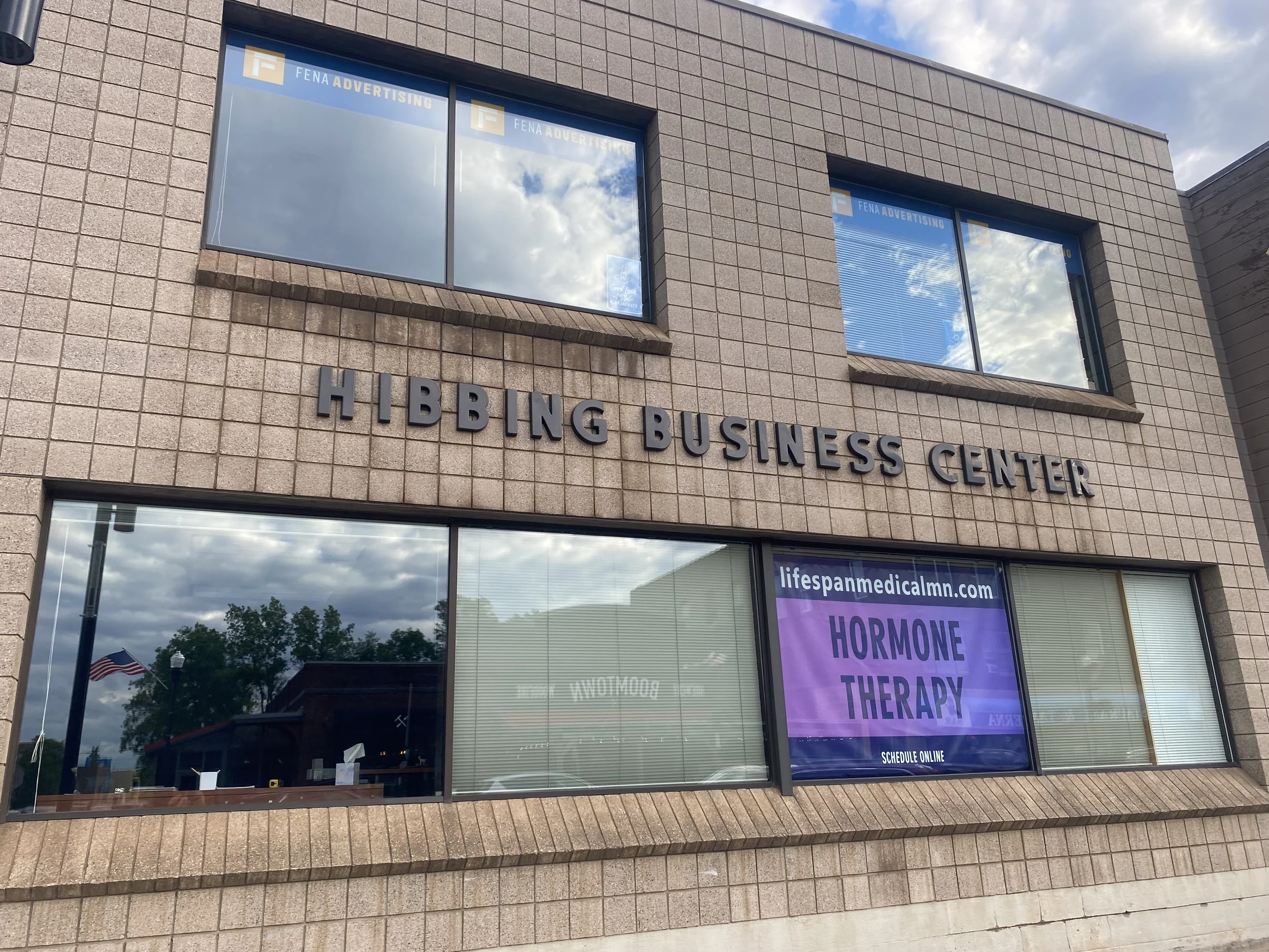 The building has a sign that reads 'HIBBING BUSINESS CENTER'. There are large windows reflecting a cloudy sky and trees. Inside, a purple banner advertises hormone therapy on Tuesday, and the website 'lifespanmedicalmn.com' is visible.
