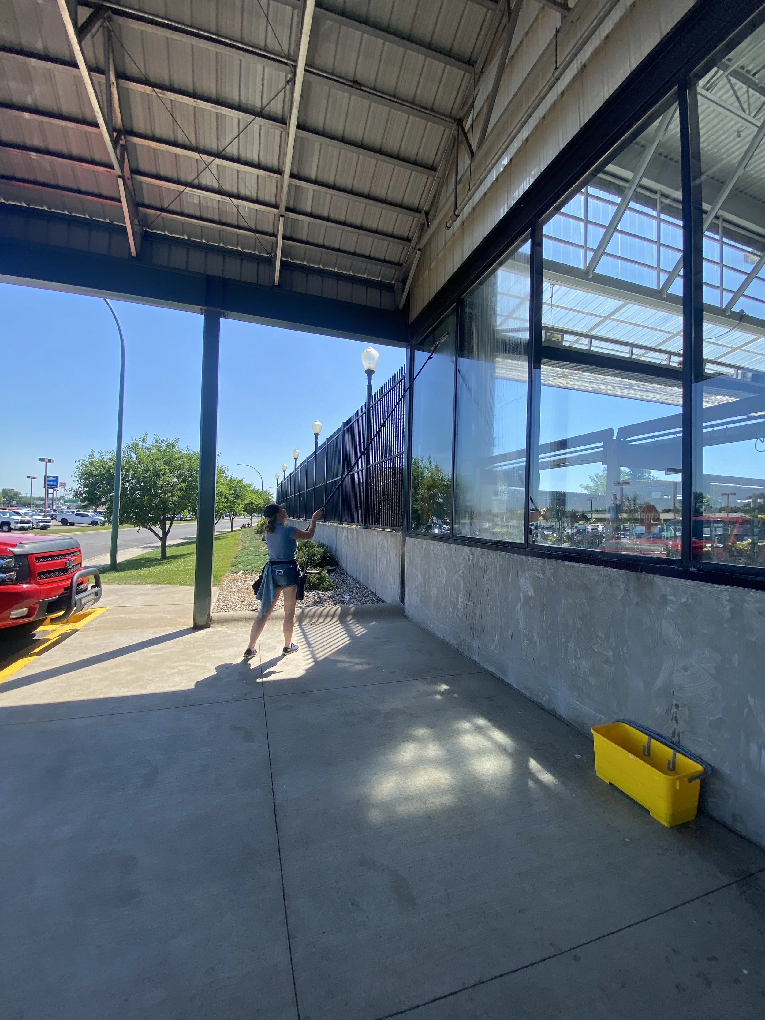 A woman cleaning the exterior window of a building with a long pole and squeegee, reflected in a large glass window on the building's wall. The scene is outdoors on a sunny day with cars and trees visible in the background.