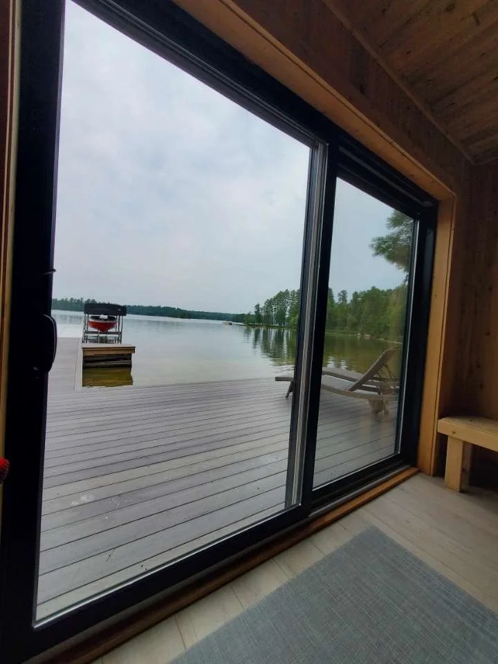 View of a serene lakeside scene through a sliding glass door, with a wooden deck, a lounge chair, and a lake surrounded by trees.