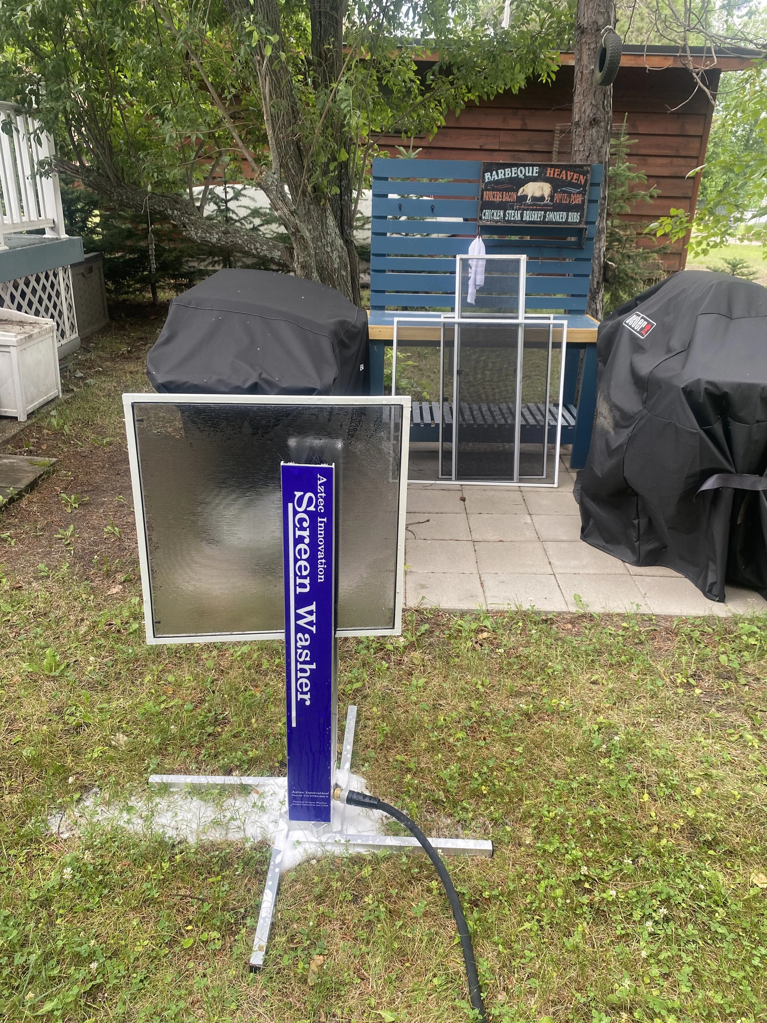 An outdoor scene with a wire-brush cleaning station, a sign indicating laser washing, and covered grills. There is a blue patio bench with a condiment sign, trees, a building in the background, and a hose connected to the cleaning station.