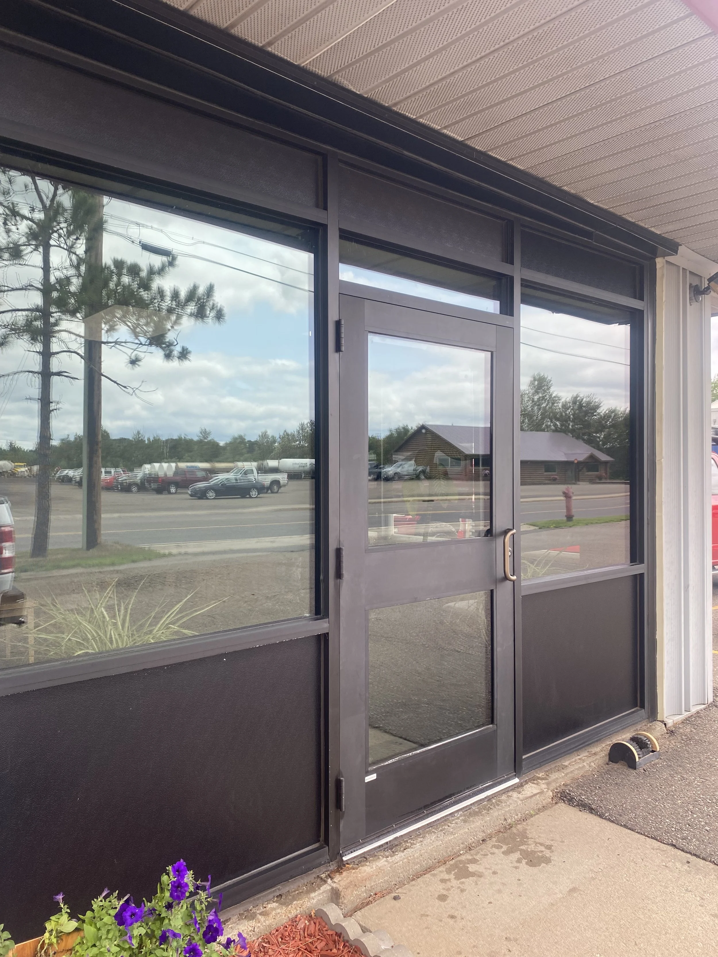 Exterior view of a storefront with large glass windows and a glass door, showing cars parked across the street, trees, and a partly cloudy sky reflected in the windows. There are potted purple flowers with green foliage on the sidewalk near the entrance.
