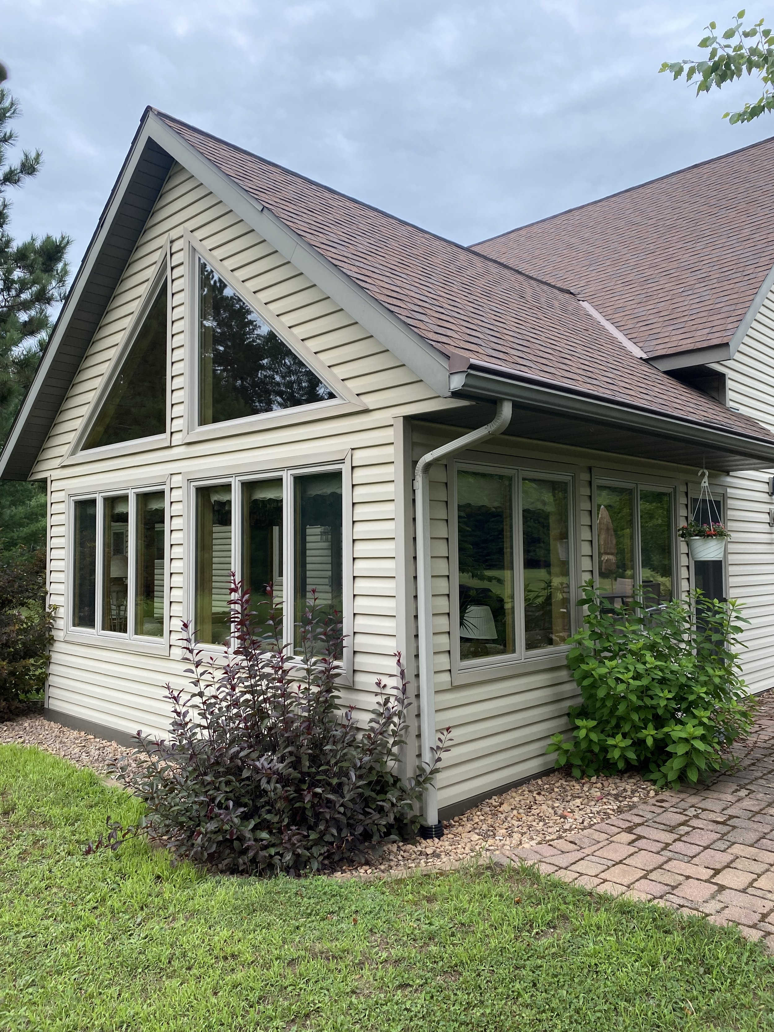 A house with beige siding, large triangular window, and a sloped roof with brown shingles. There are bushes and grass in the yard, and a small brick pathway. window have been professionally cleaned