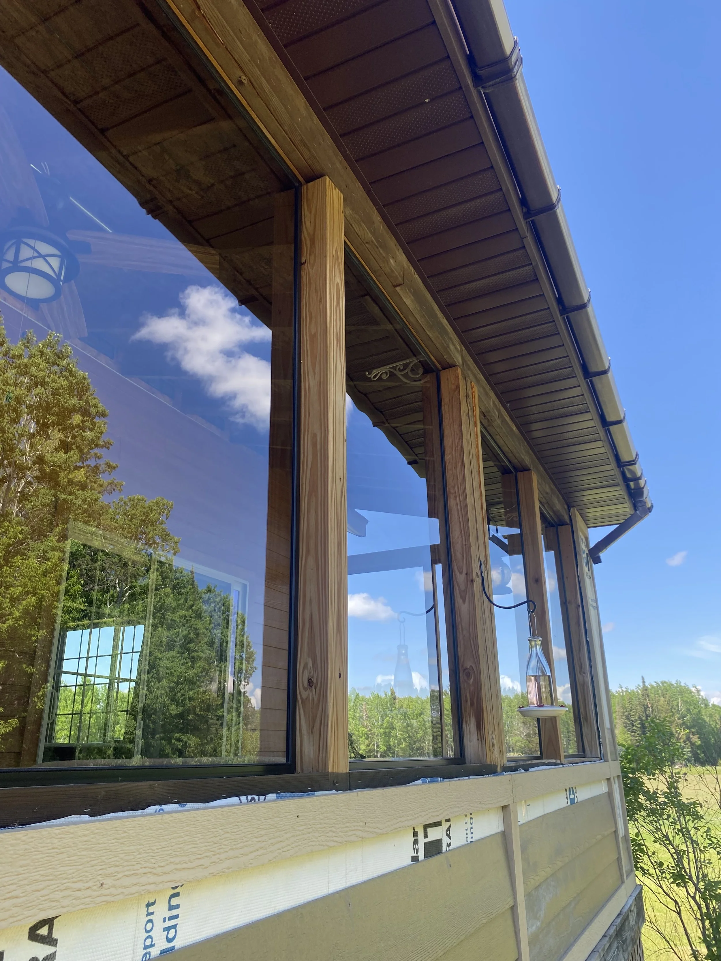Partially constructed house exterior with large window reflecting trees and blue sky, wooden framing, and building materials in the foreground during daytime.