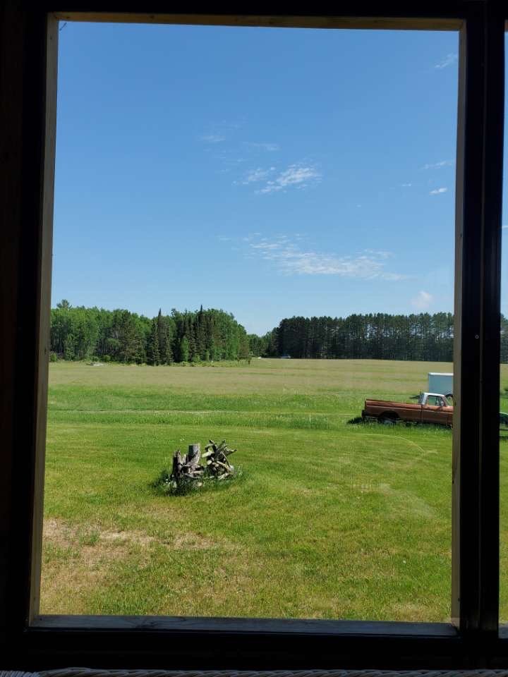 View through a window showing a green lawn, a tree stump, a red truck, and a white trailer under a blue sky with some clouds.