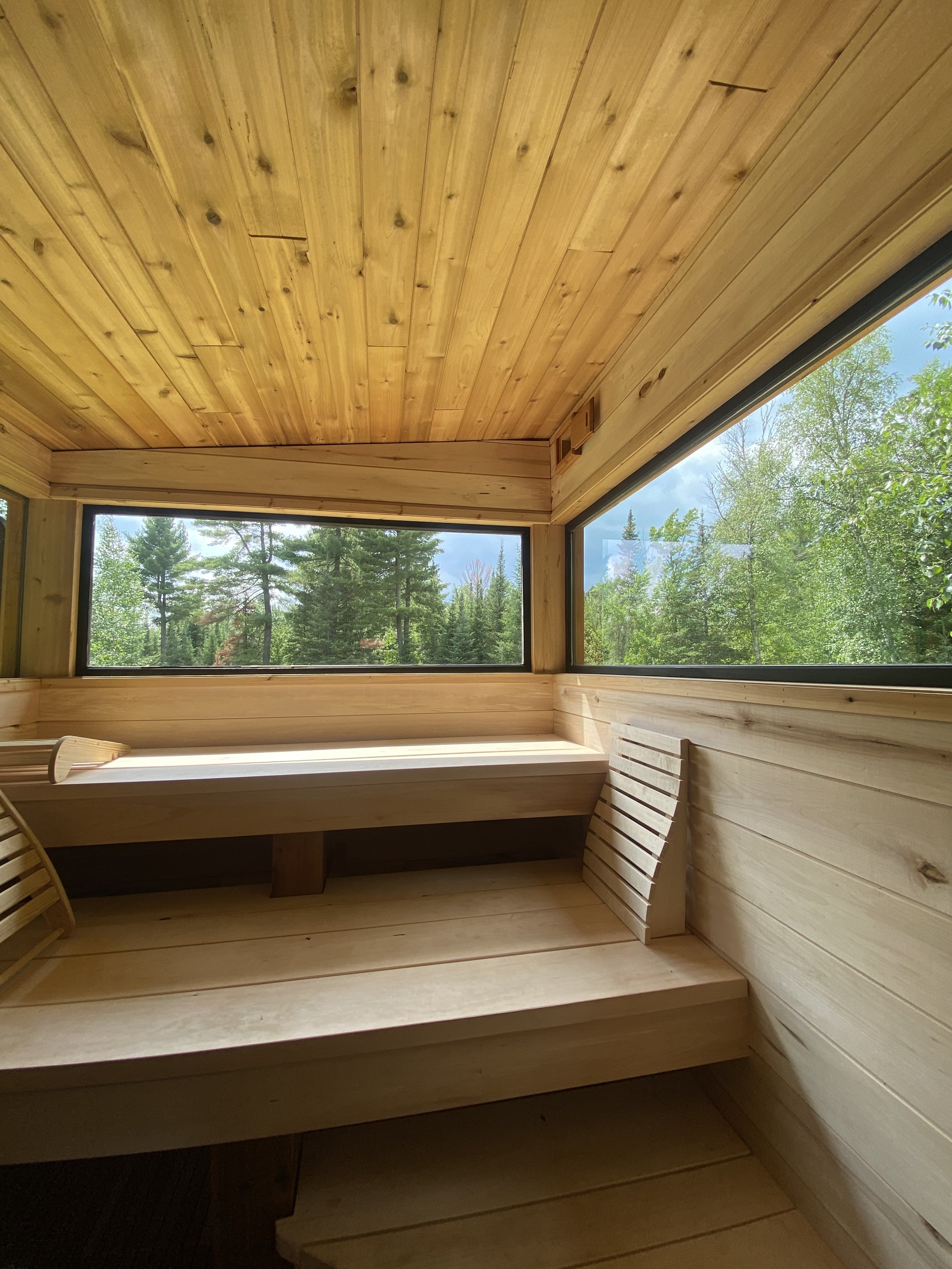 A cozy wooden sauna room with large windows showing trees and a blue sky outside. The room has a wooden bench and slatted backrests inside.