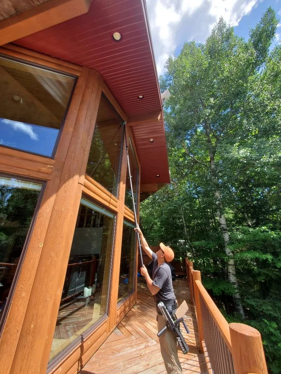 A person cleaning the exterior window of a wooden house using a long cleaning tool. The house has large glass windows, a red roof overhang, and is surrounded by green trees under a partly cloudy sky.