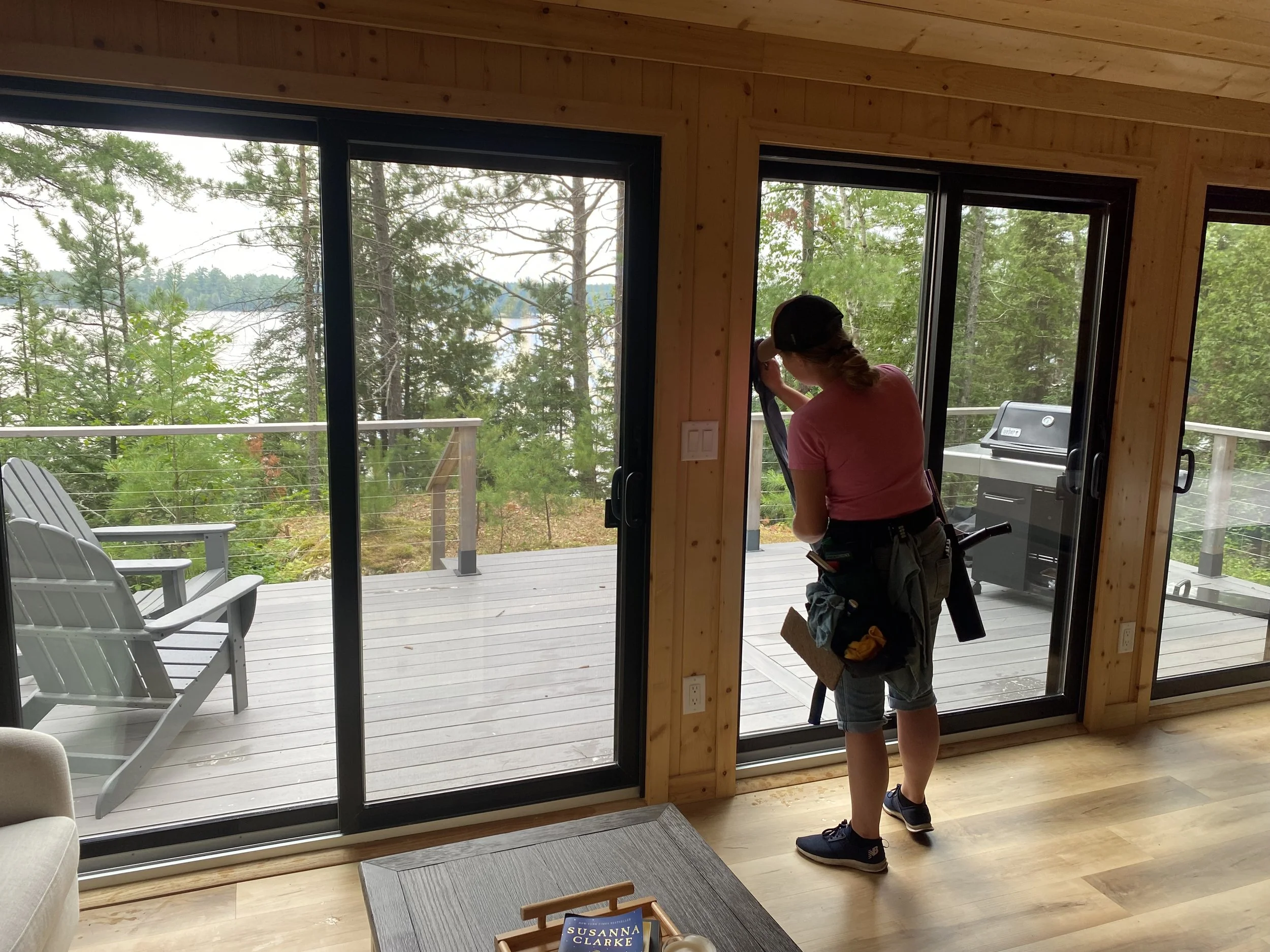 A person cleaning glass door outside of a wooden house, with a deck and trees in a wooded area behind